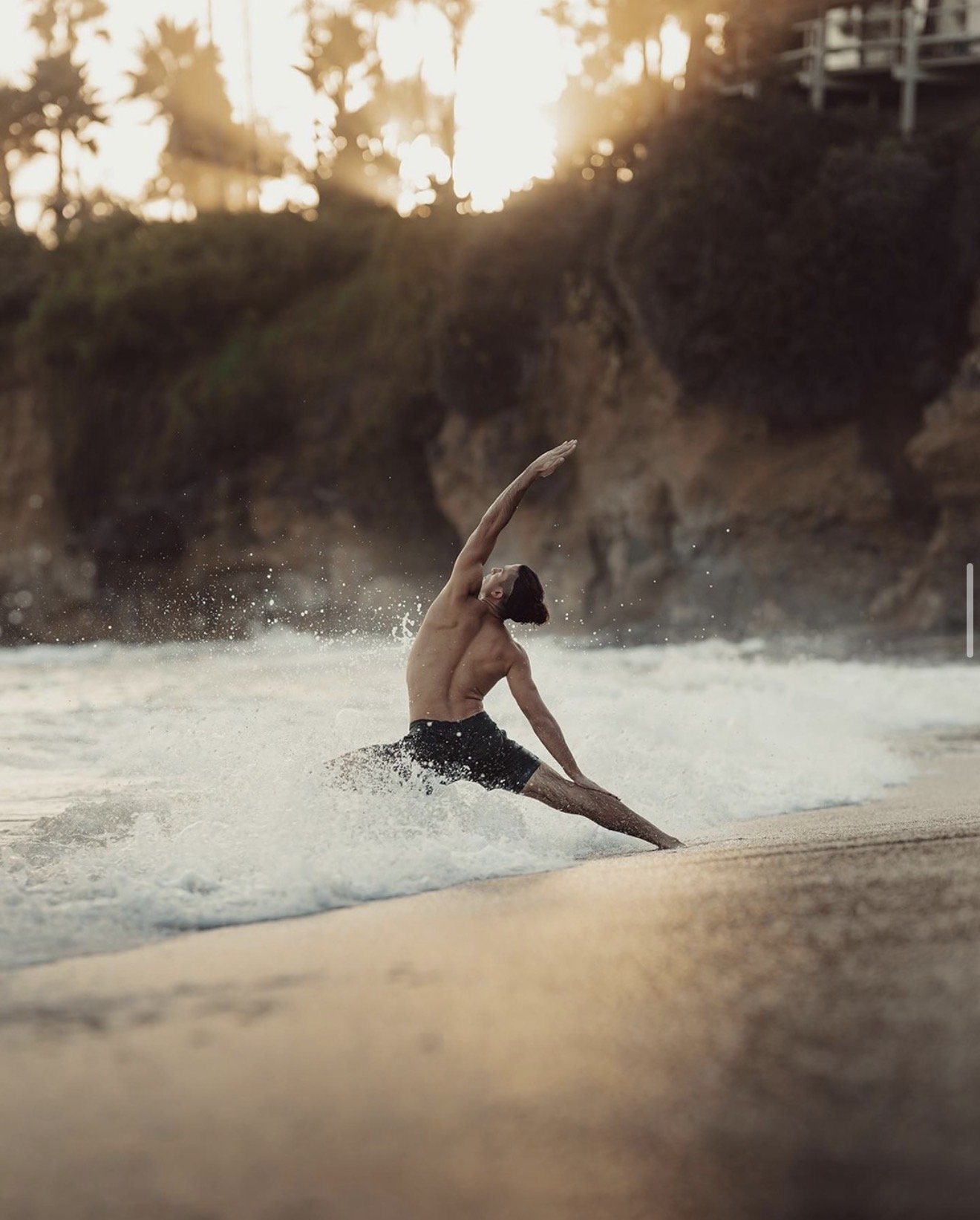 A man practicing yoga on the beach during sunset, with waves splashing around him and rocky cliffs in the background.