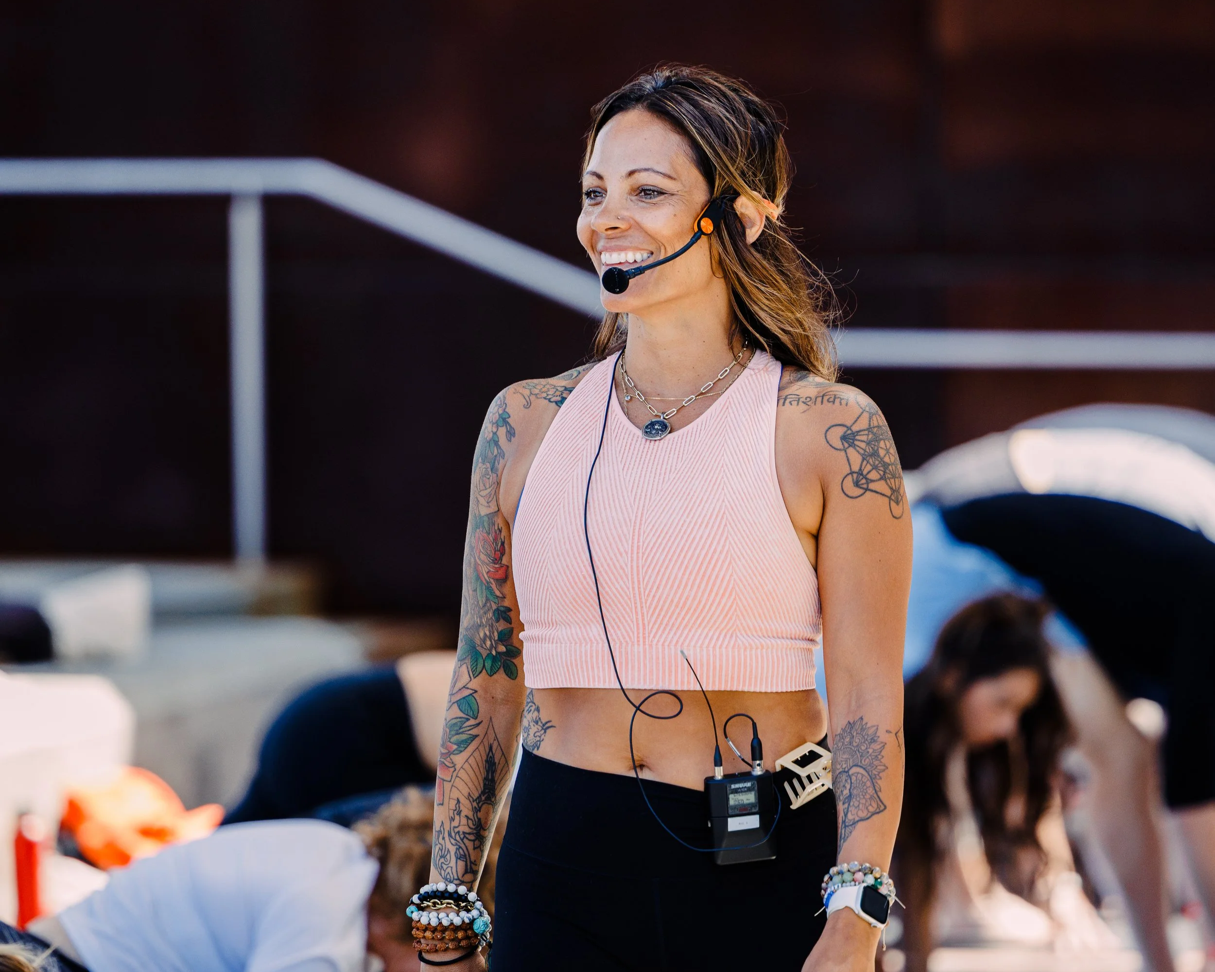 A woman with tattoos on her arms, wearing a pink sleeveless crop top, several bracelets, and a necklace, is smiling while wearing a headset microphone at an outdoor event.