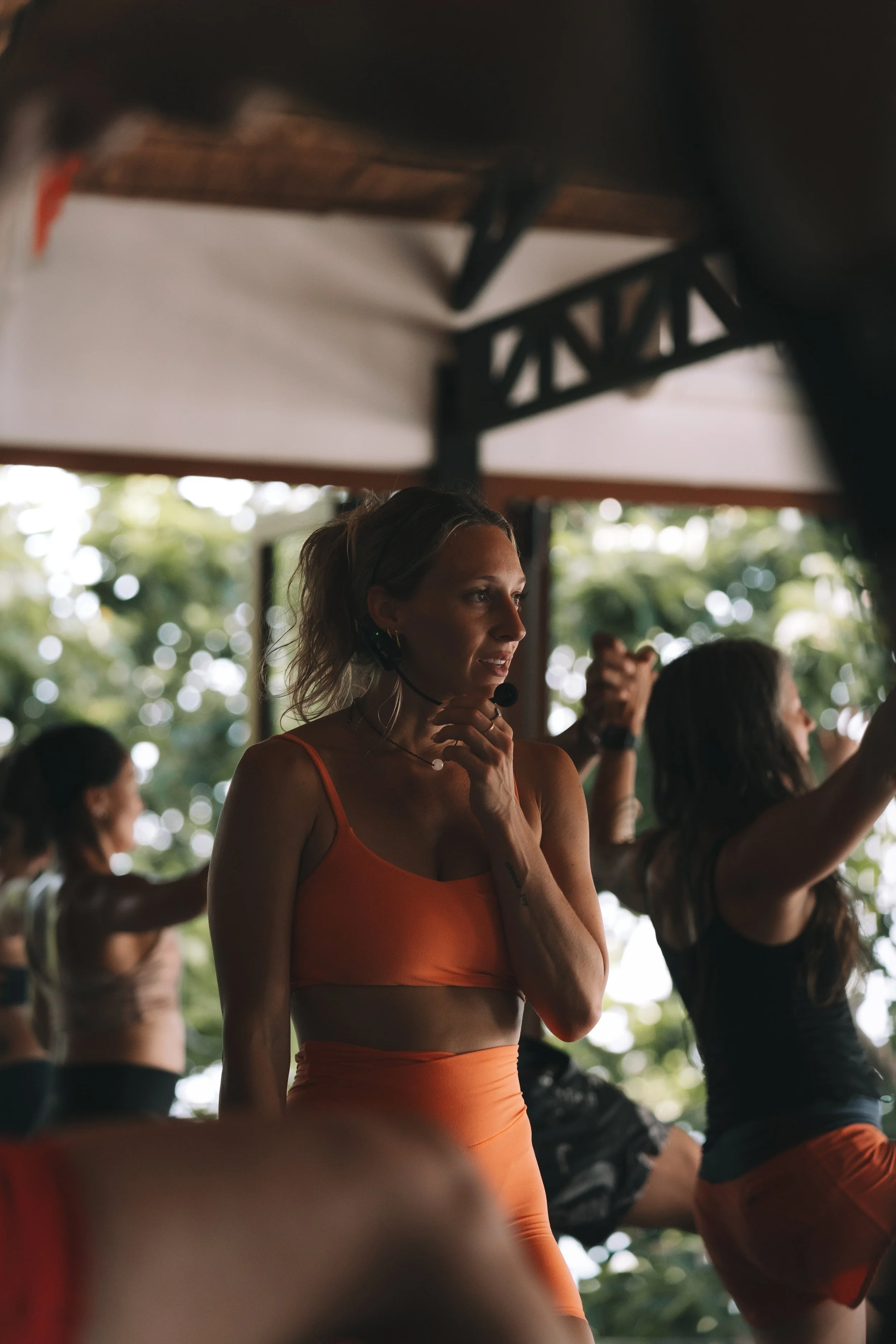 A woman with blonde hair in a yoga or exercise class, wearing an orange sports bra and leggings, with a microphone headset, in a wooden studio with trees outside.