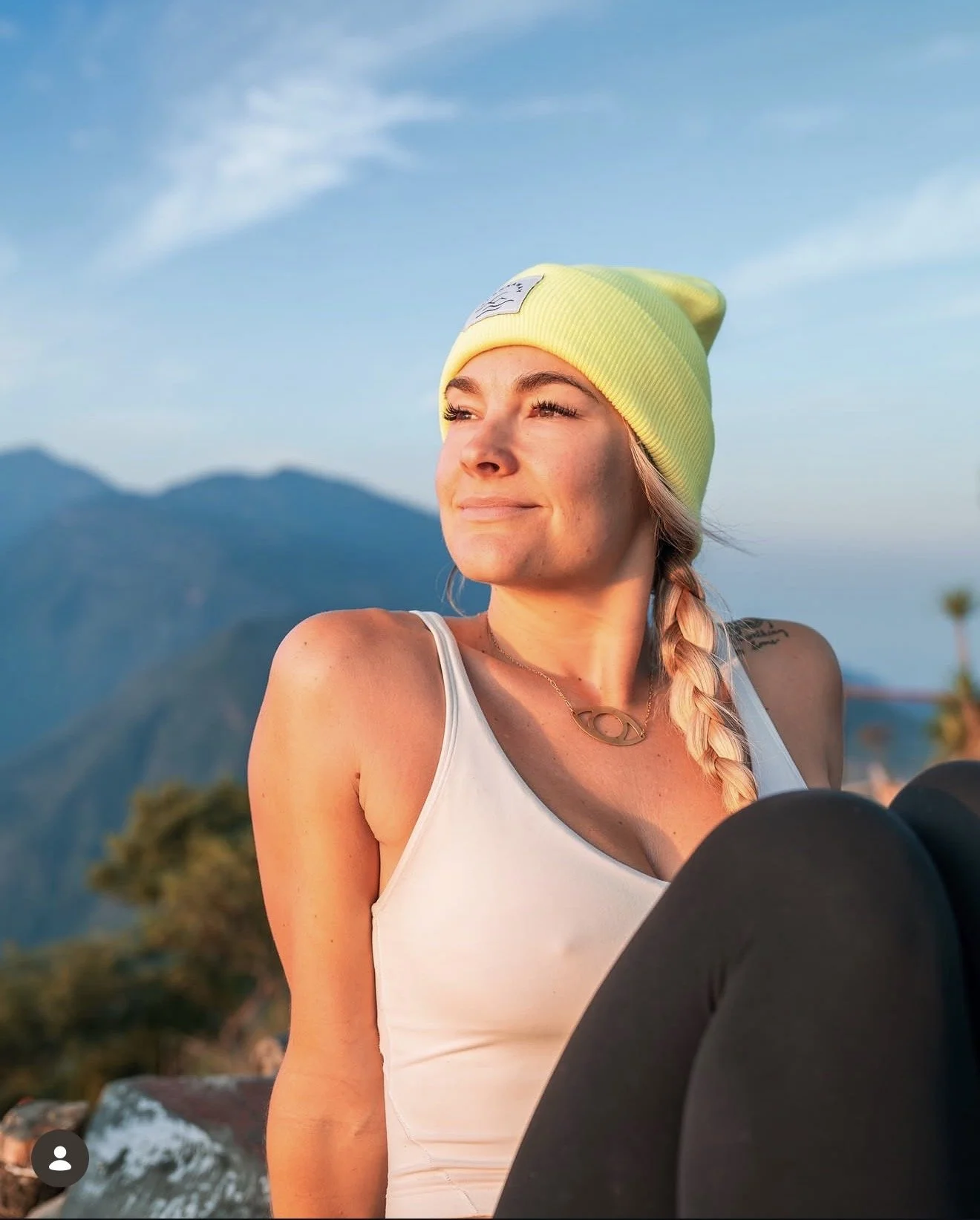 A woman with a blonde braid wearing a yellow beanie and white tank top, sitting outdoors with mountains in the background, enjoying the sunset