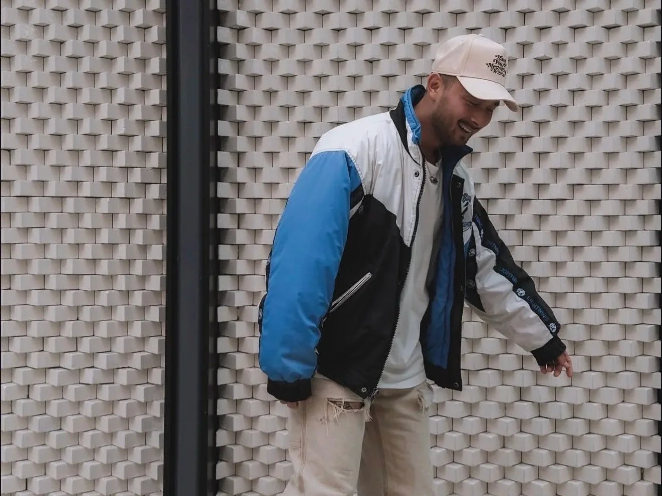 Man wearing a beige cap, black, white, and blue North Face jacket, and beige pants, standing against a textured wall, smiling and looking down.