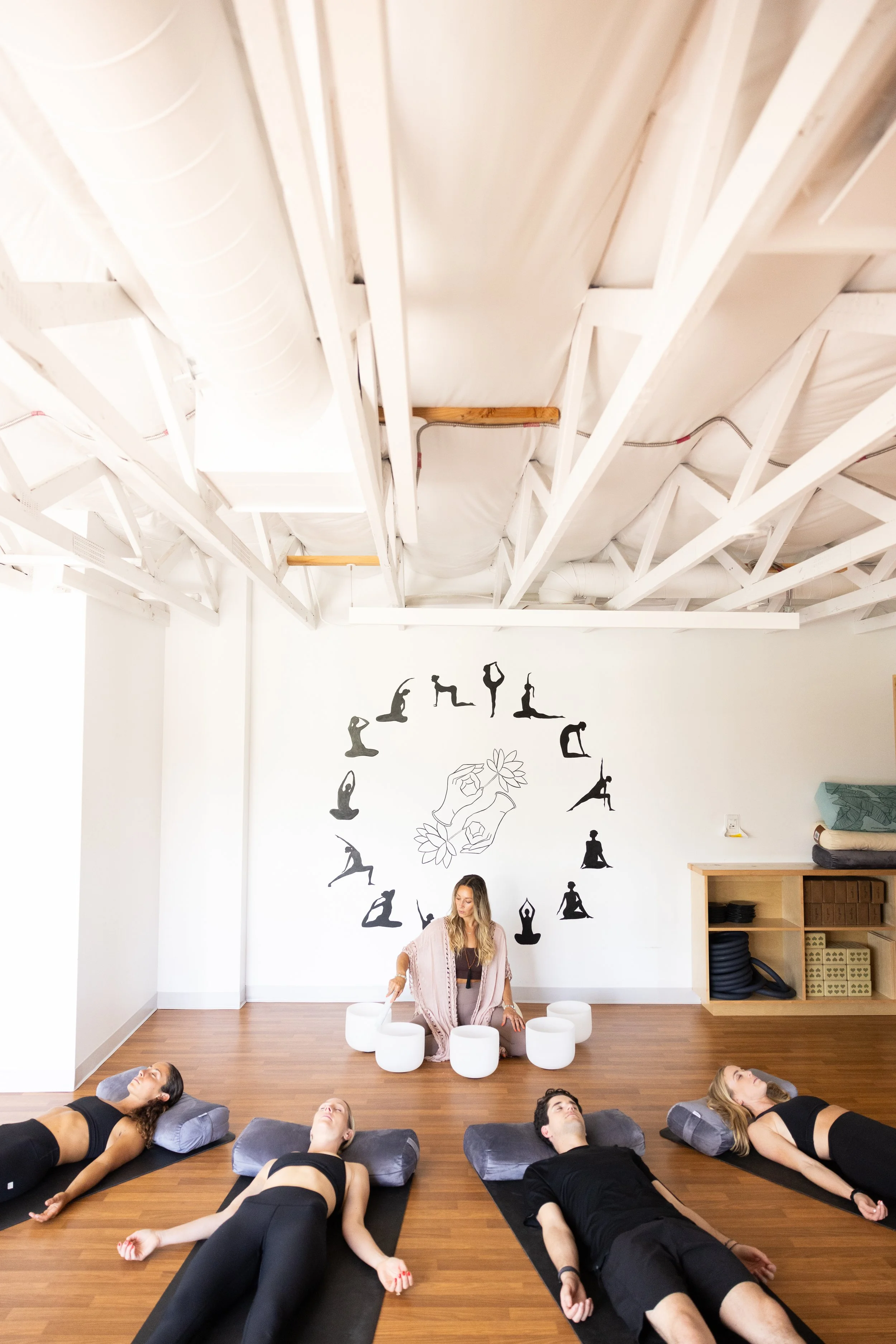 Yoga class with five people lying on mats and pillows, a woman leading meditation seated cross-legged on a fitness ball, yoga silhouettes and a lotus design on the wall
