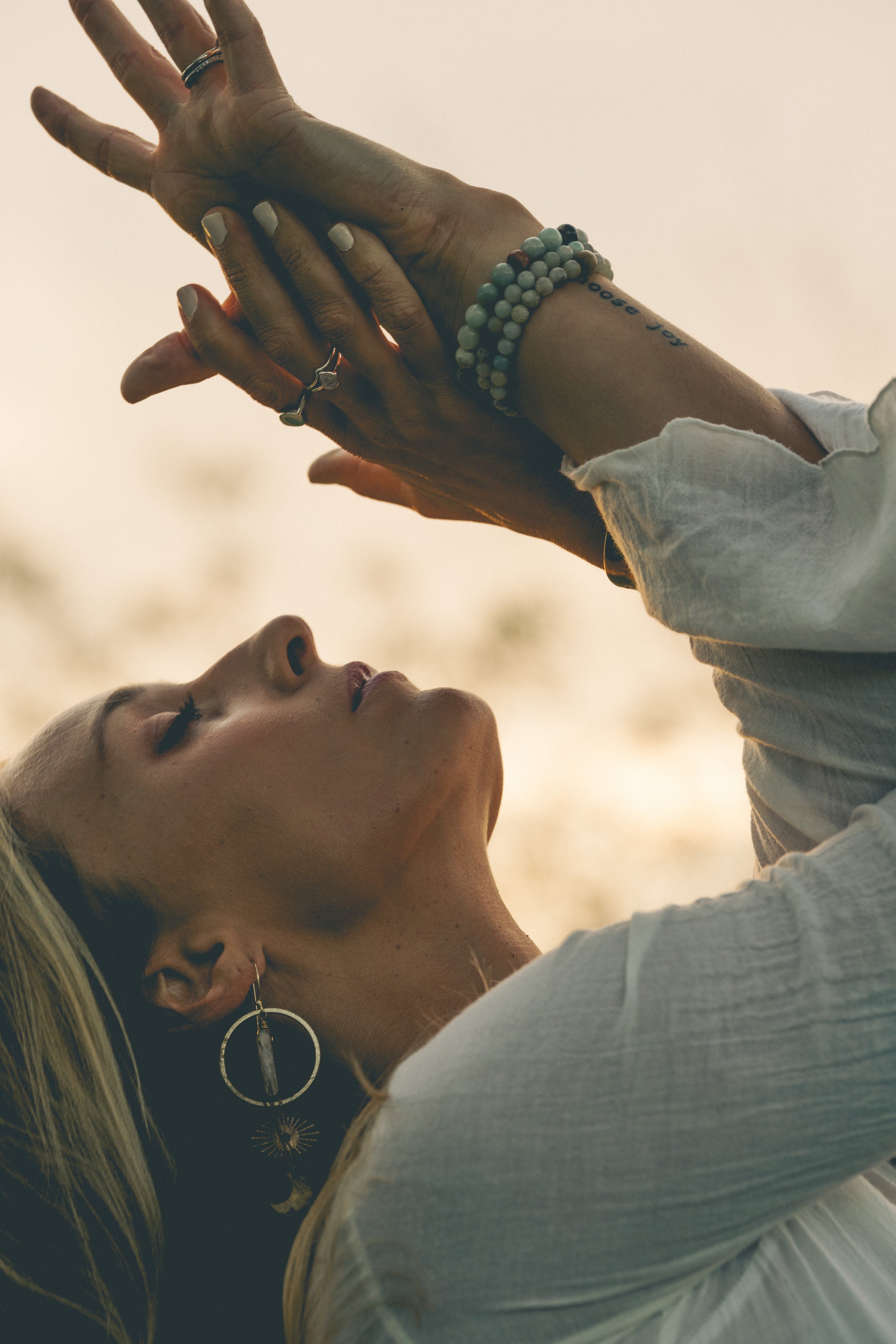 A person with platinum blonde hair and multiple earrings has closed eyes and is lying with their head tilted back. Their hands, adorned with rings and bracelets, are gently touching their face and shoulder. The background is blurred and neutral-color