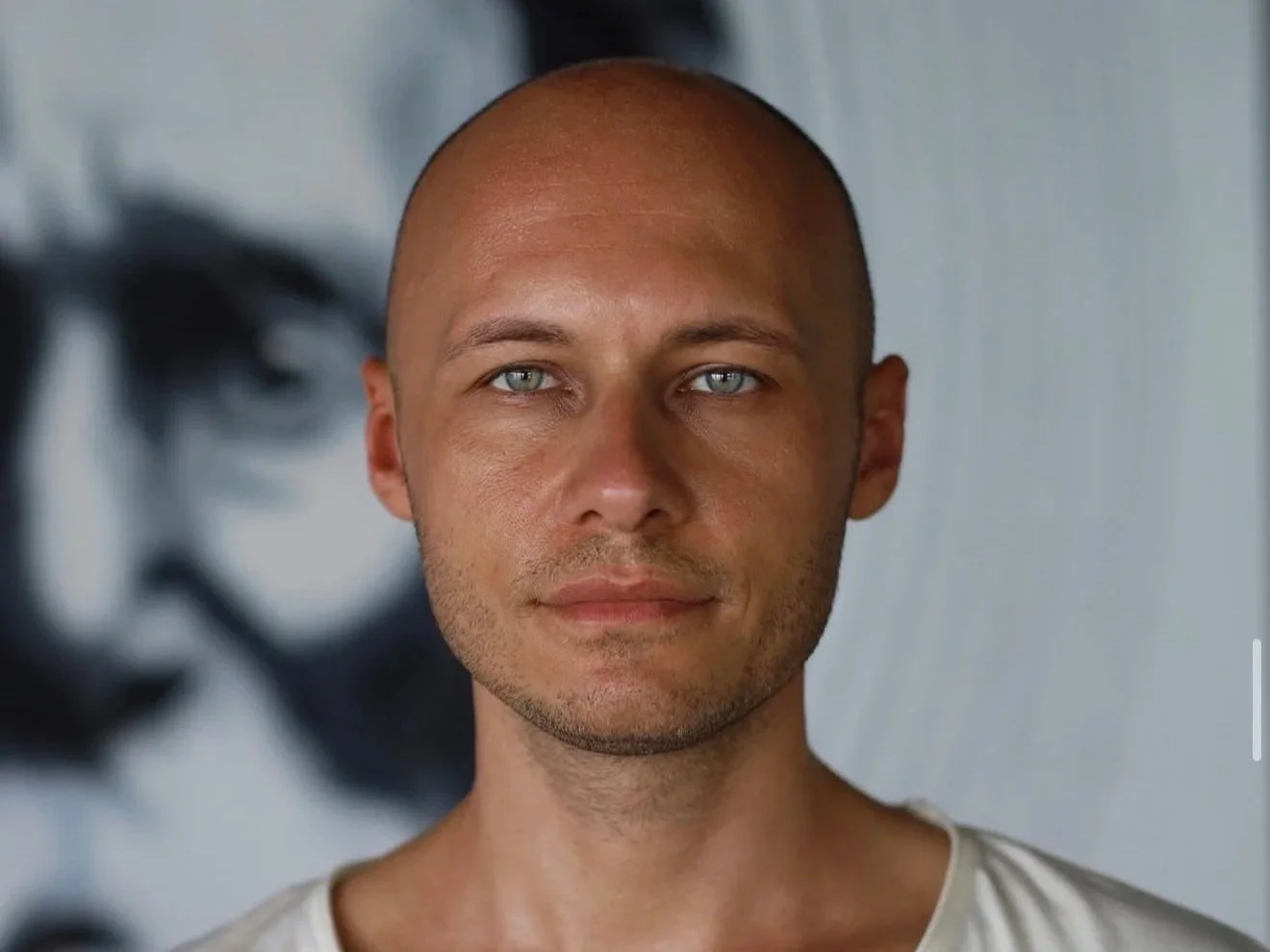 Close-up of a man with light skin, a shaved head, and blue eyes, looking directly at the camera, with a black and white face artwork in the background.