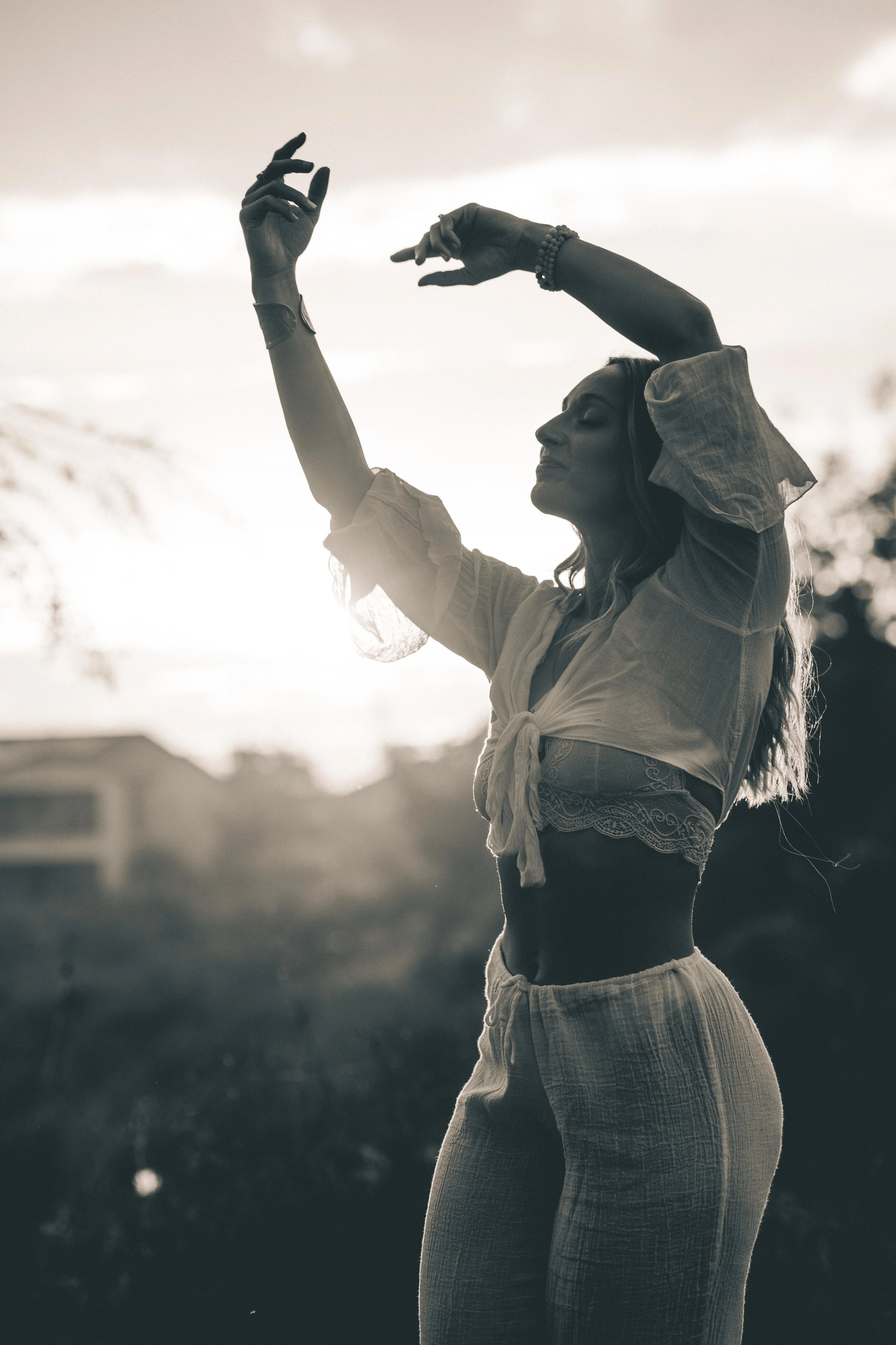 A woman in a relaxed pose with her eyes closed, wearing a short-sleeved shirt and pants, standing outdoors with a blurred background and cloudy sky, captured in black and white.