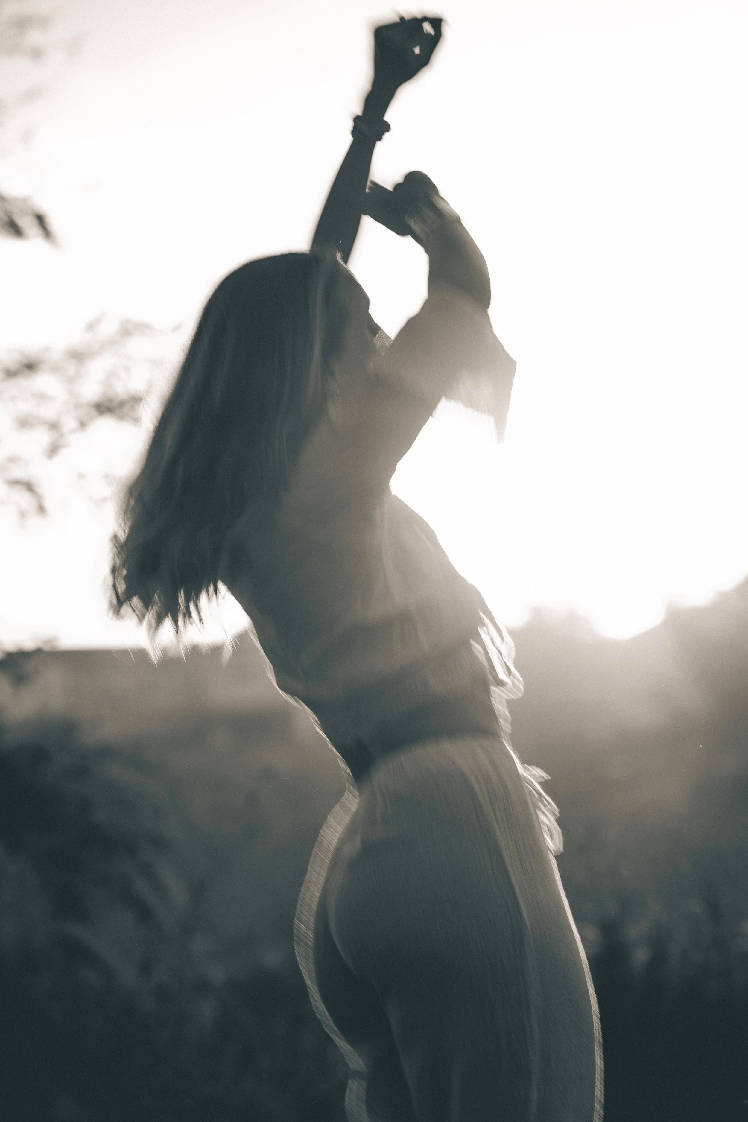 A woman casually dressed, wearing a sweater and baggy jeans, reaching upward with one arm, standing outdoors during daytime with the sun behind her, creating a silhouette effect.