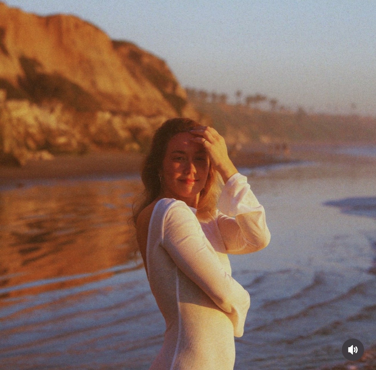 A woman standing on a beach during sunset, with her hand on her forehead, smiling at the camera. The background features ocean waves and rocky cliffs.
