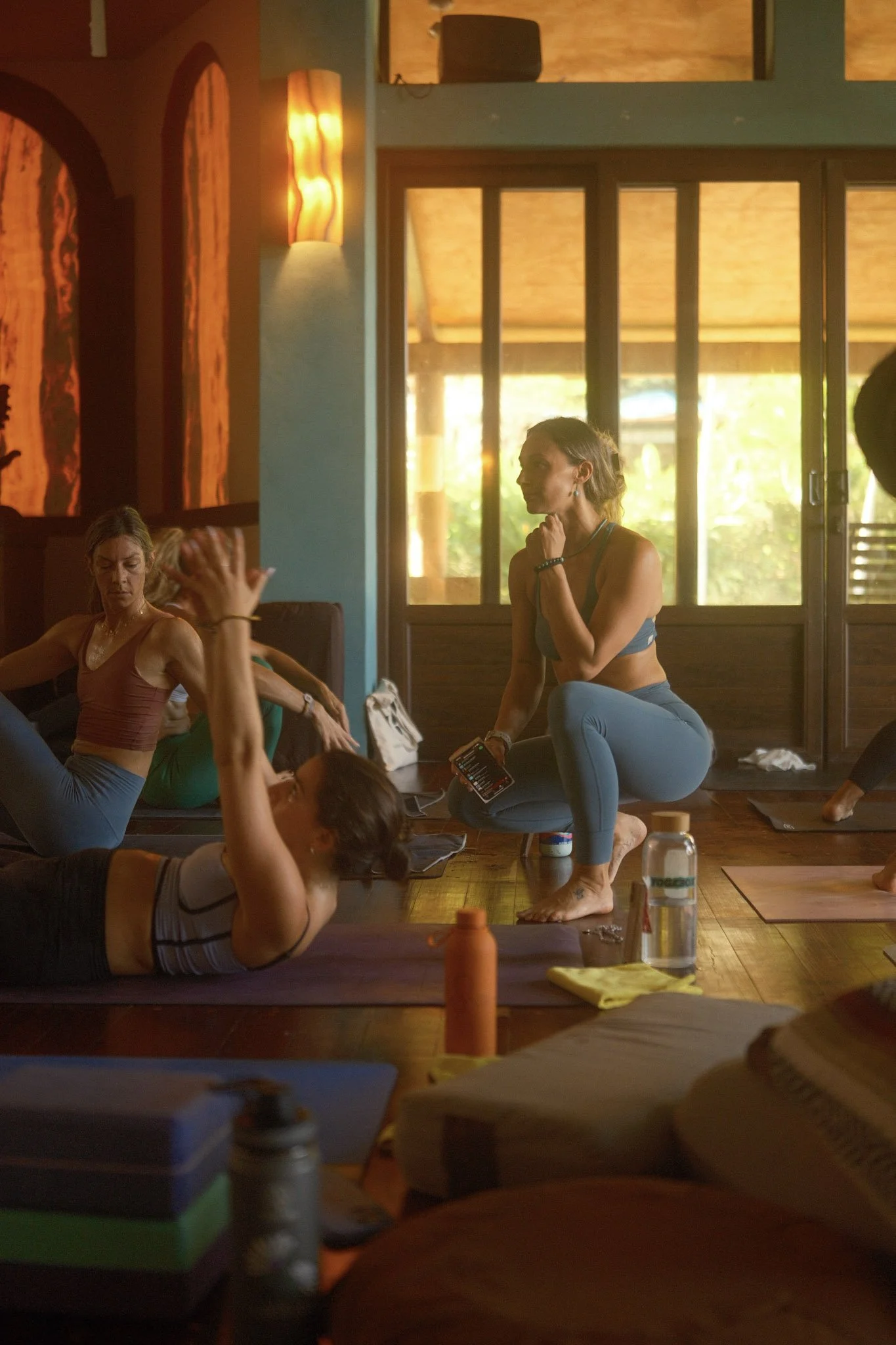 Women participating in a yoga or meditation class inside a wooden-floored room with large windows, sunlight streaming in, some seated or kneeling on mats, engaged in relaxation or stretching exercises.