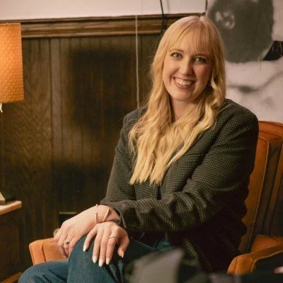 A woman with long blonde hair, smiling, sitting in a wooden-paneled room.
