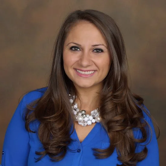 Portrait of a smiling woman with long brown hair, wearing a royal blue blouse and a large pearl necklace, against a neutral brown background.