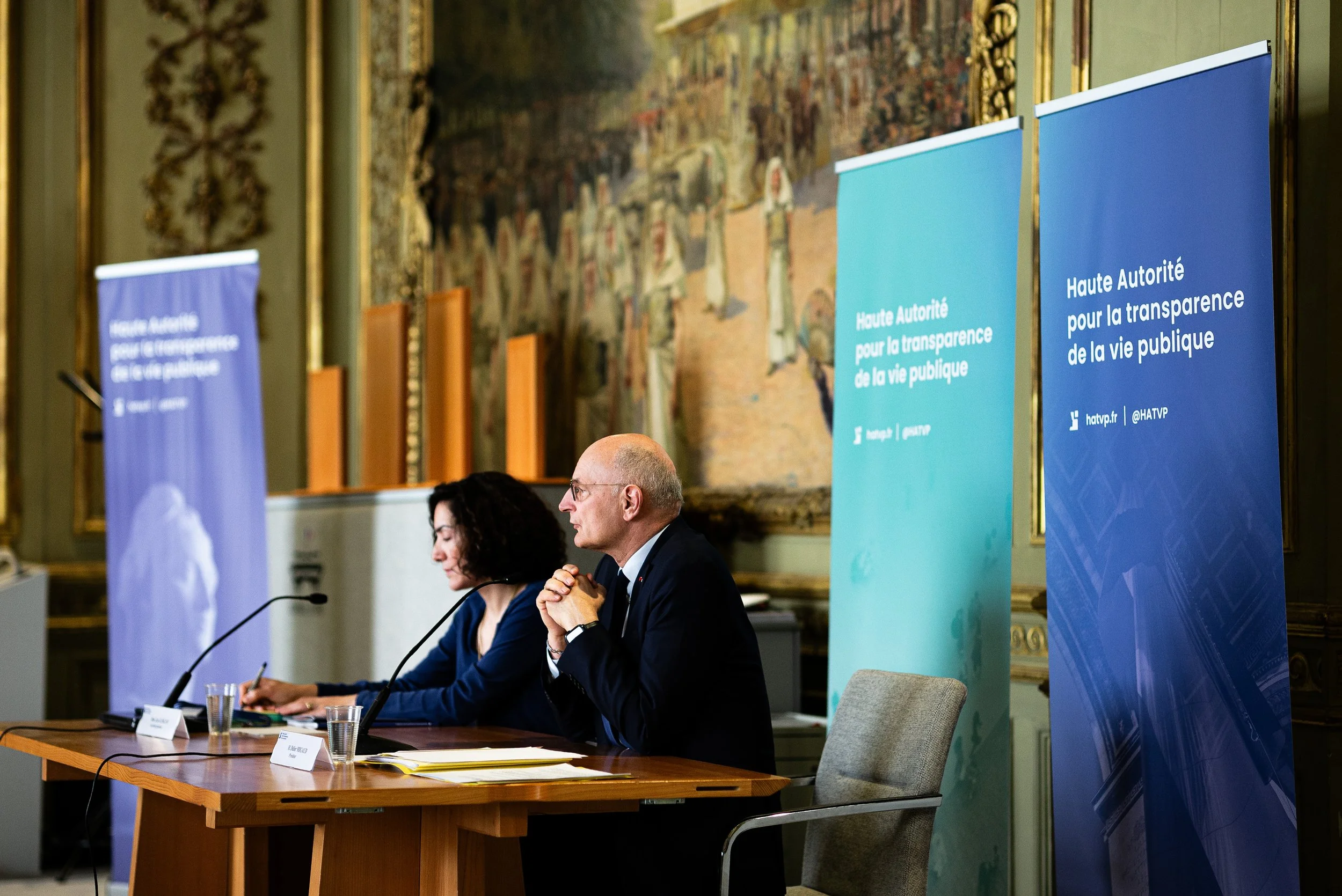Deux personnes assises à une table lors d'une conférence, avec des banderoles bleues et vertes en arrière-plan, dans un décor élégant avec des peintures et des moulures dorées.