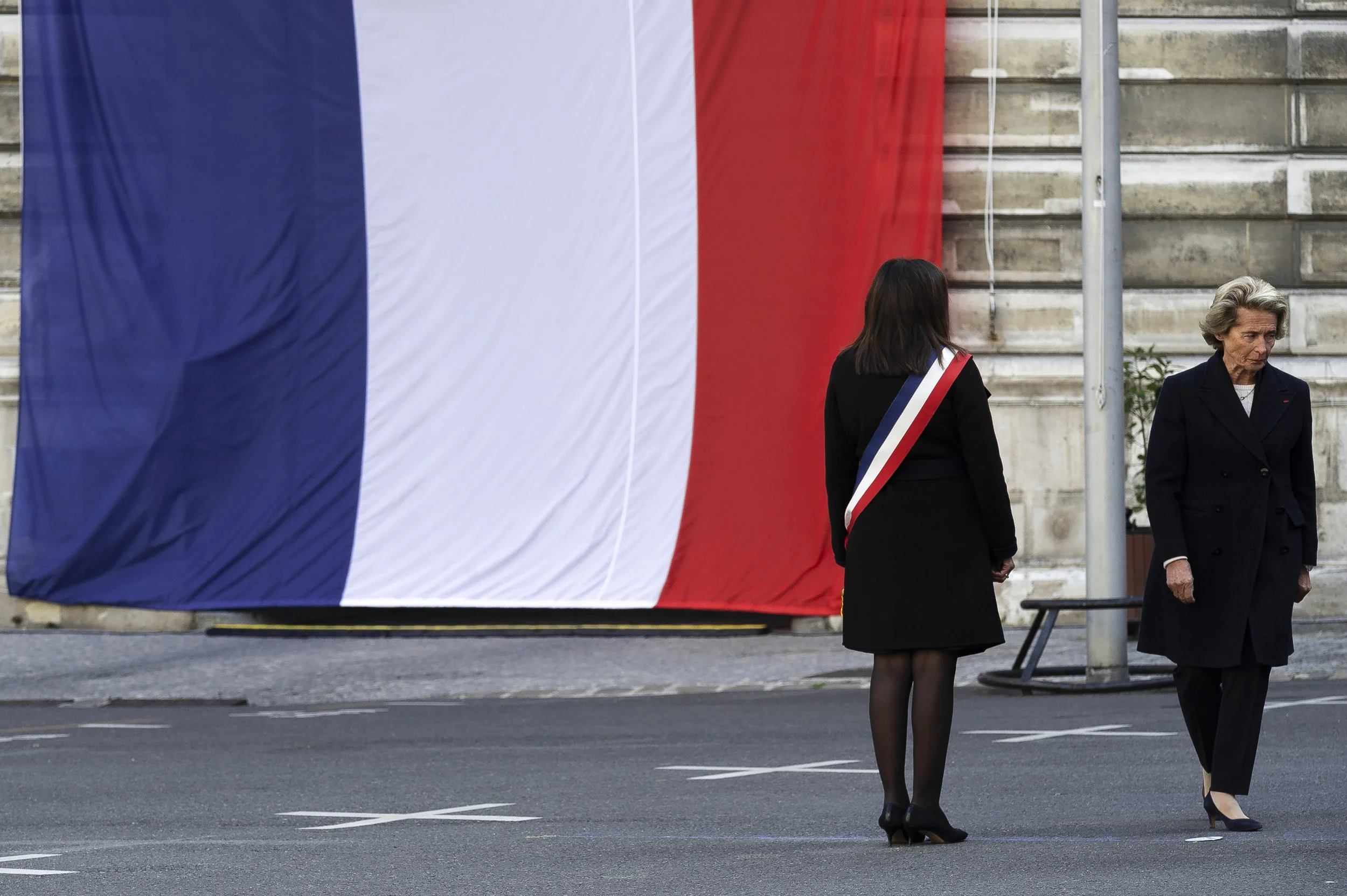 Deux femmes en manteaux noirs et talons hauts, dont une porte une écharpe tricolore (bleu, blanc, rouge), se tiennent devant un grand drapeau tricolore français suspendu derrière elles, devant un bâtiment en pierre.
