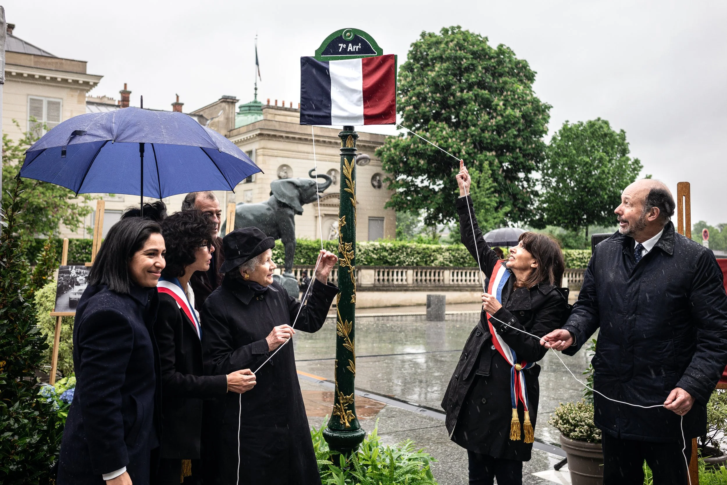 Groupe de personnes en vêtements noirs, utilisant une corde pour dévoiler un drapeau français lors d'un événement sous la pluie à l'extérieur, avec un bâtiment historique, une statue de chien, des arbres et des pots de fleurs en arrière-plan.