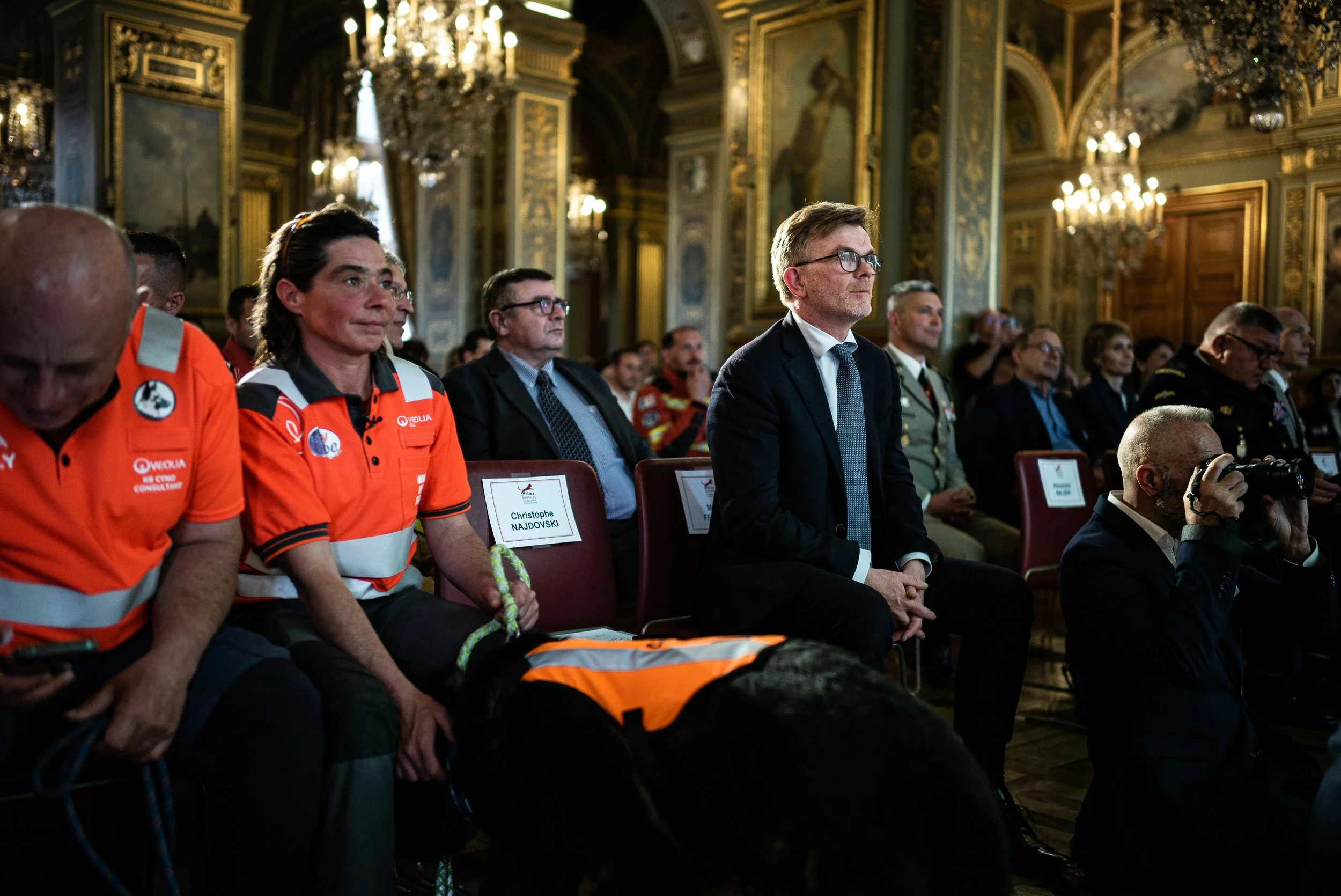 Audience lors d'une cérémonie officielle dans une salle ornée de dorures et de lustres. Parmi les personnes, un homme en costume sombre, une femme en uniforme orange, et un chien en gilet orange, assis dans la première rangée.