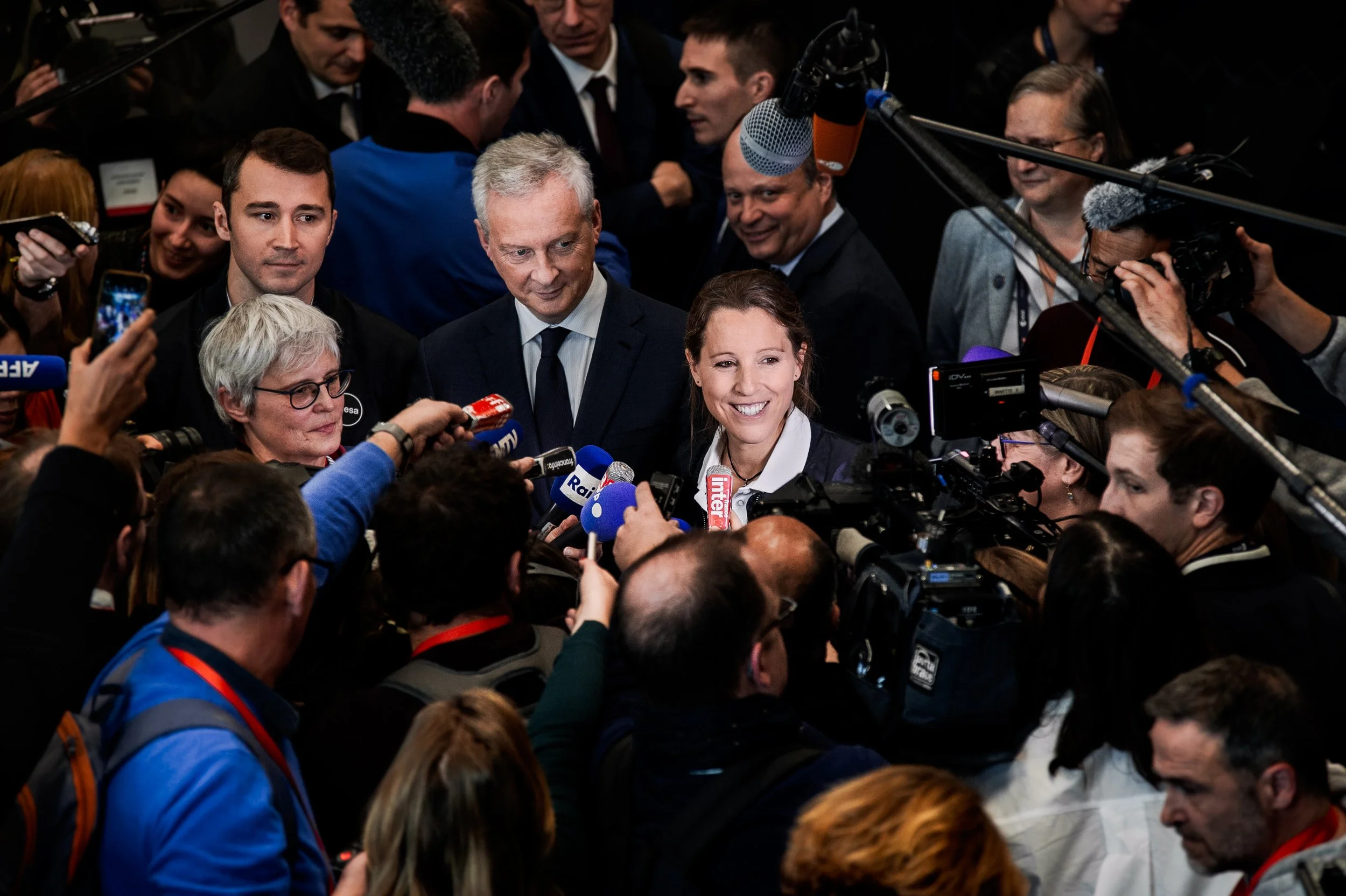 Une femme souriante entourée de journalistes et de caméramans lors d'une conférence de presse ou d'un événement médiatique.