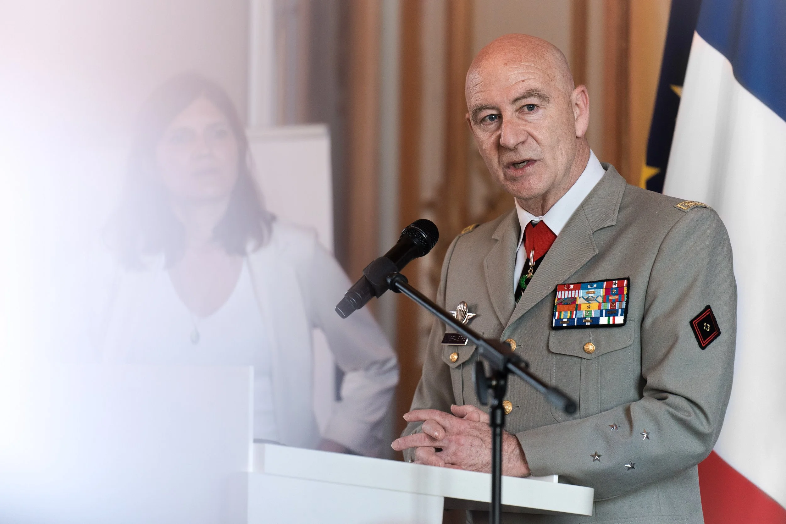 Homme en uniforme militaire parlant à un micro lors d'une cérémonie, avec un drapeau français en arrière-plan.