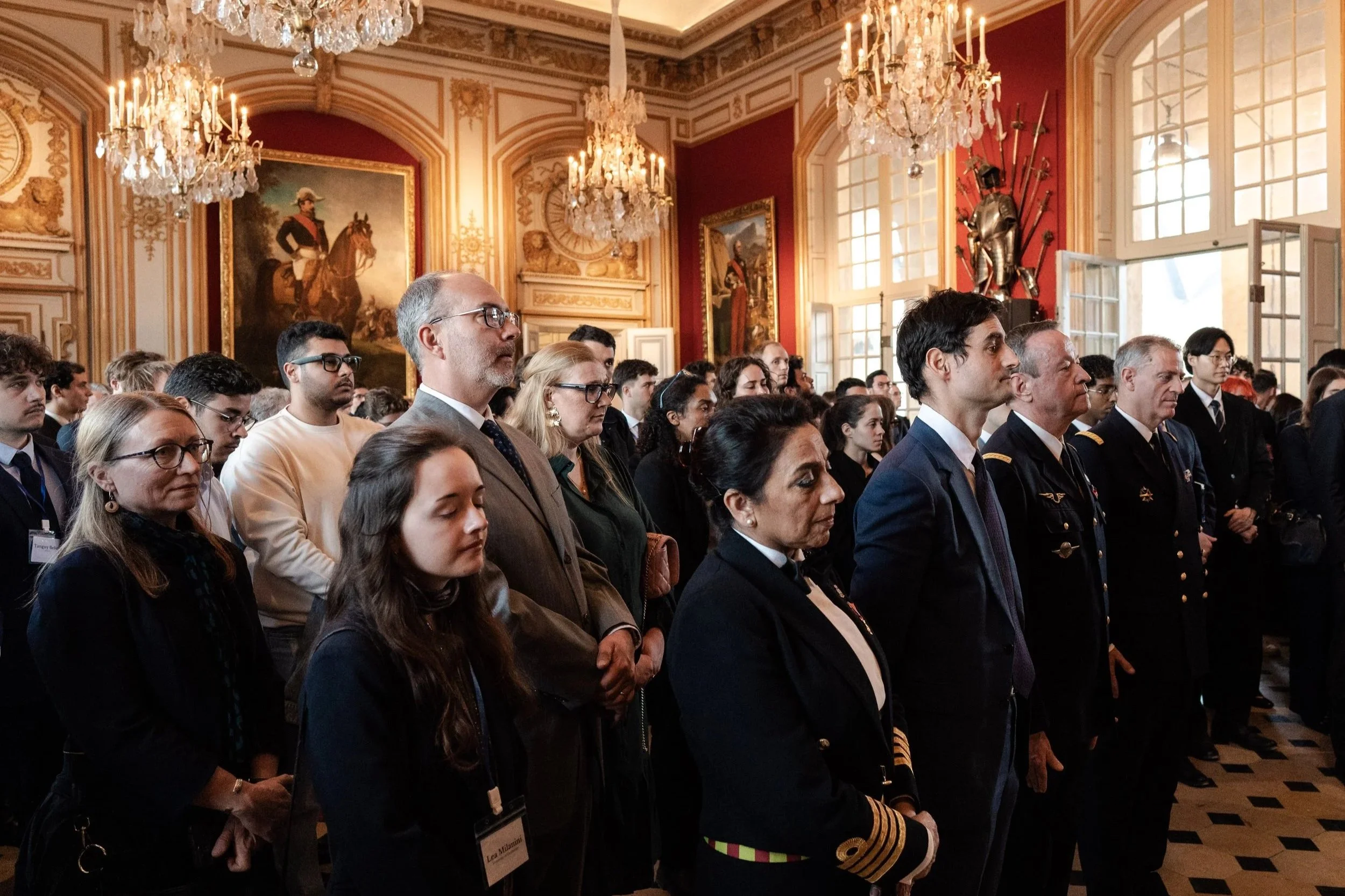 Un groupe de personnes à un événement officiel dans une salle richement décorée avec des chandeliers, des peintures et des armures anciennes, face à l'avant-plan.