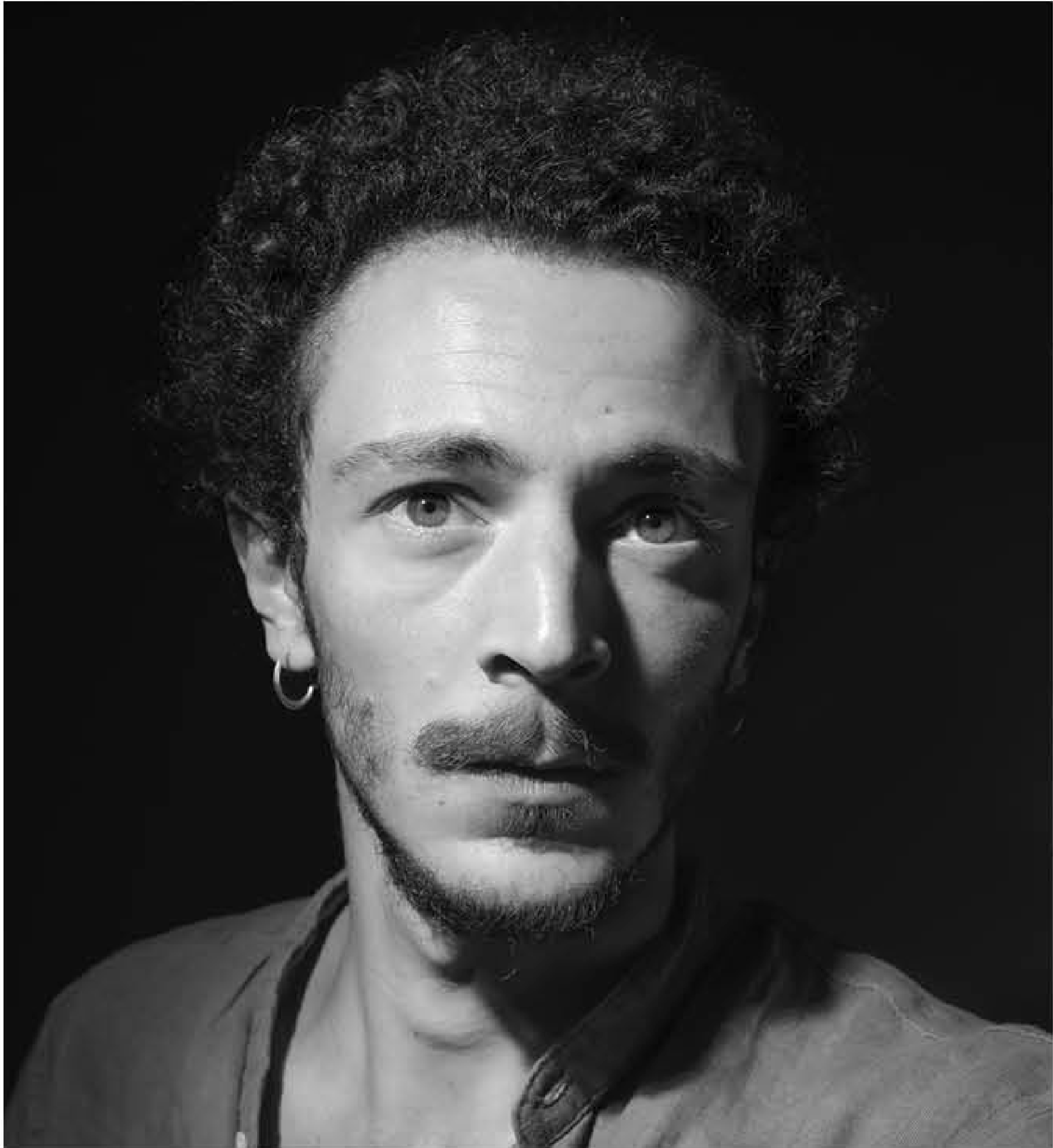 Portrait en noir et blanc d'un homme avec des cheveux bouclés, une boucle d'oreille à l'oreille gauche, et un tatouage sur le cou, portant une veste ouverte.