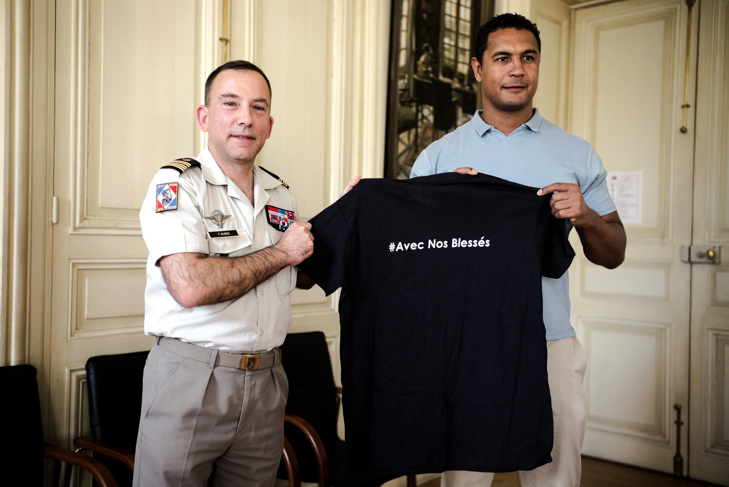 Deux hommes, un en uniforme militaire et un autre en chemise bleue, tiennent ensemble un T-shirt noir avec l'inscription '#Avec Nos Blessés' dans une pièce avec des murs clairs et des chaises en bois.