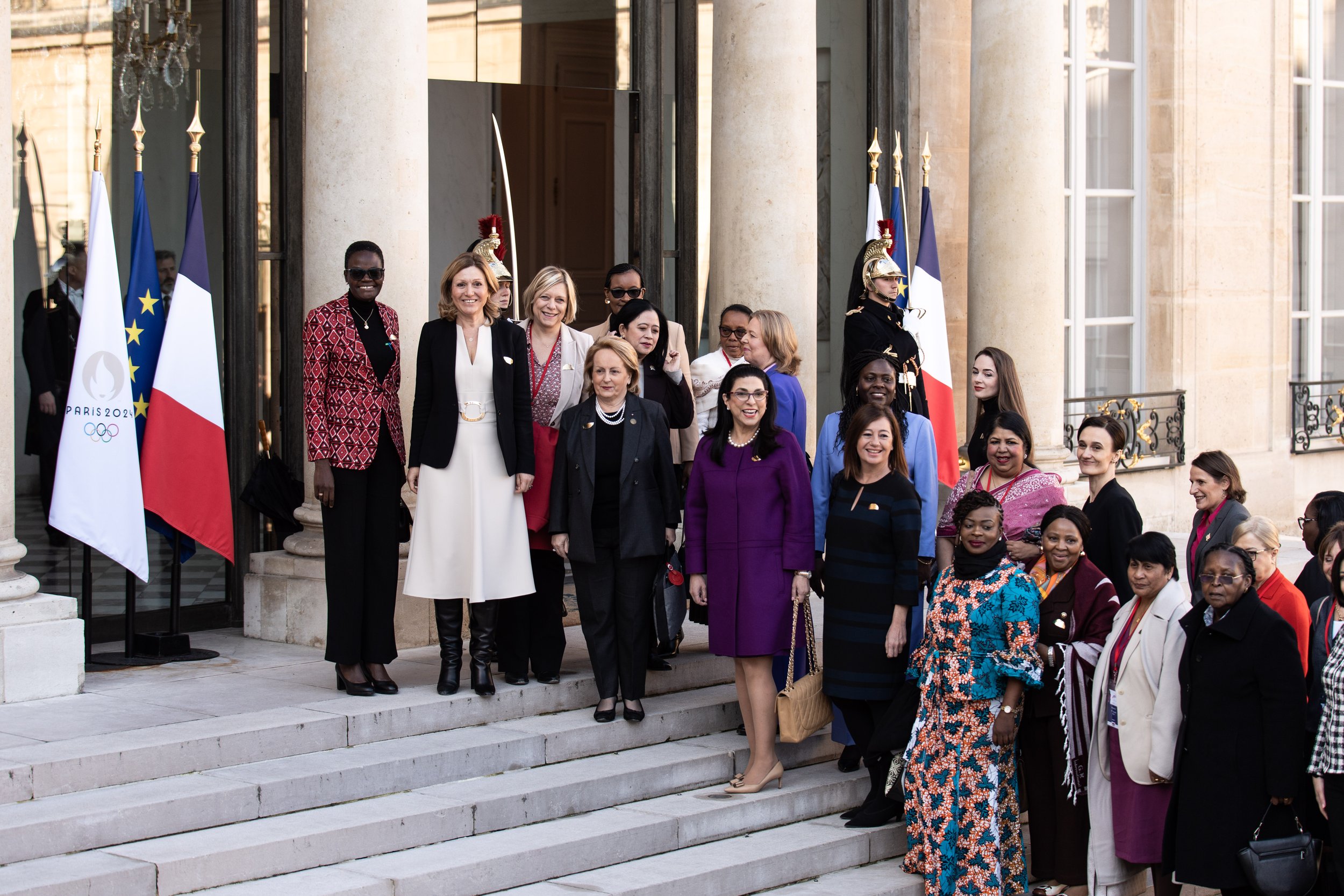 Groupe de femmes posant sur les marches devant une façade architecturale, avec drapeaux français, européen et Jeux Olympiques de Paris 2024, lors d'un événement officiel en France.