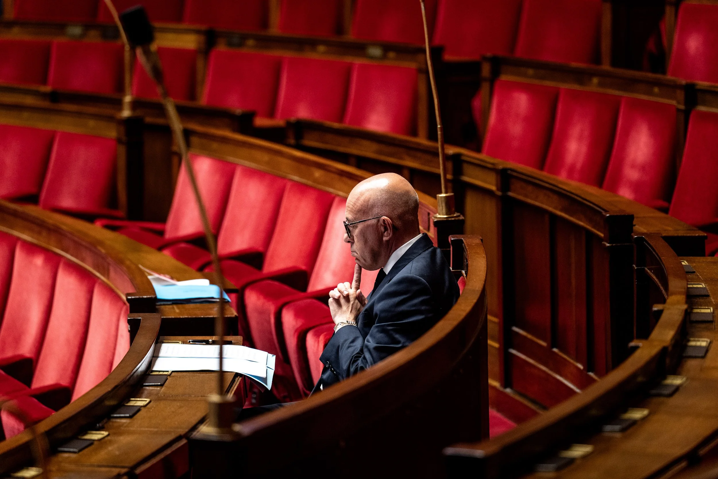 Un homme assis seul dans un siège de l'assemblée avec des sièges rouges, probablement dans un parlement ou une salle de réunion officielle, en train de réfléchir ou de se concentrer.