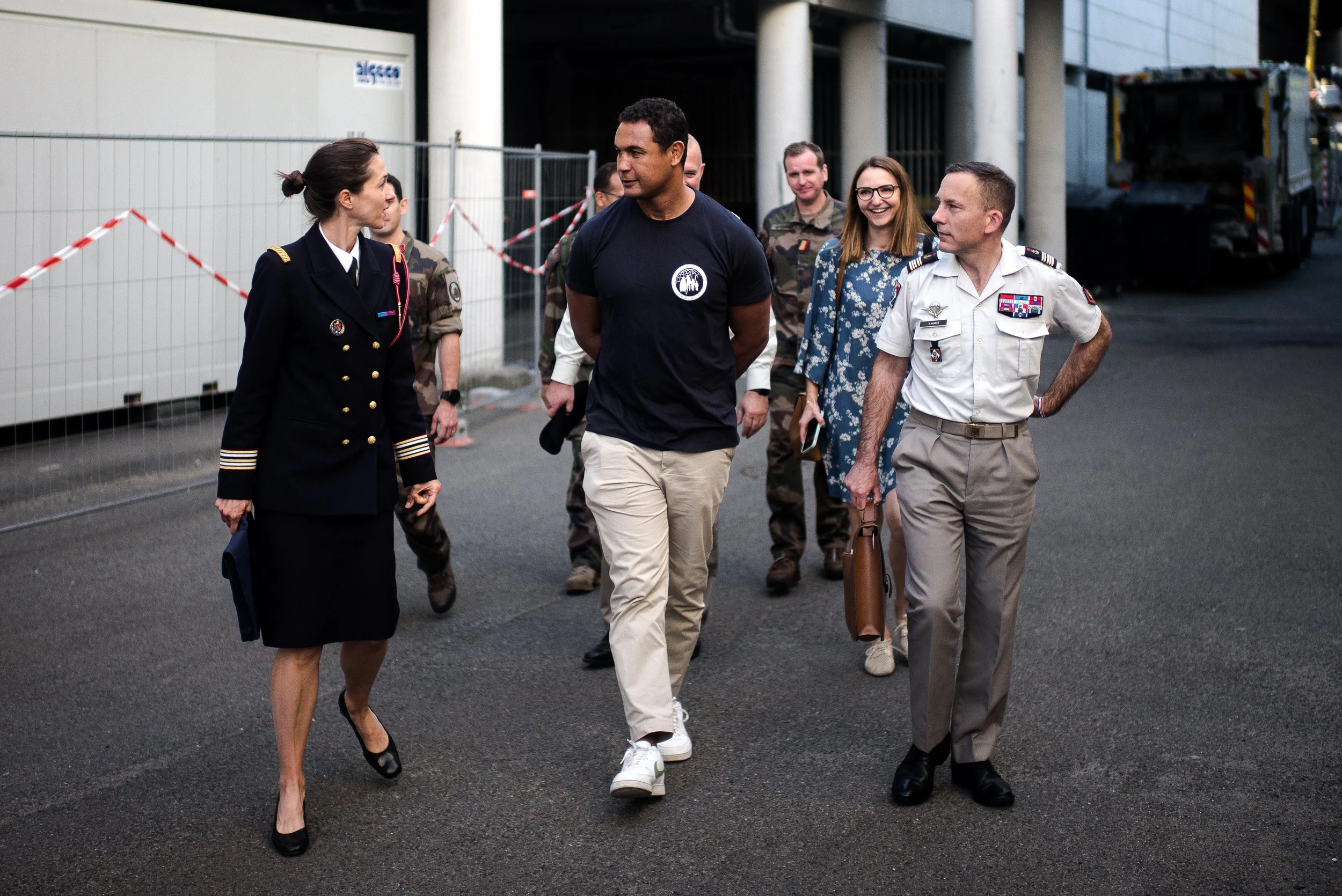 Groupe de personnes en uniforme militaire et civil marchant et discutant dehors près d'une barrière de sécurité.