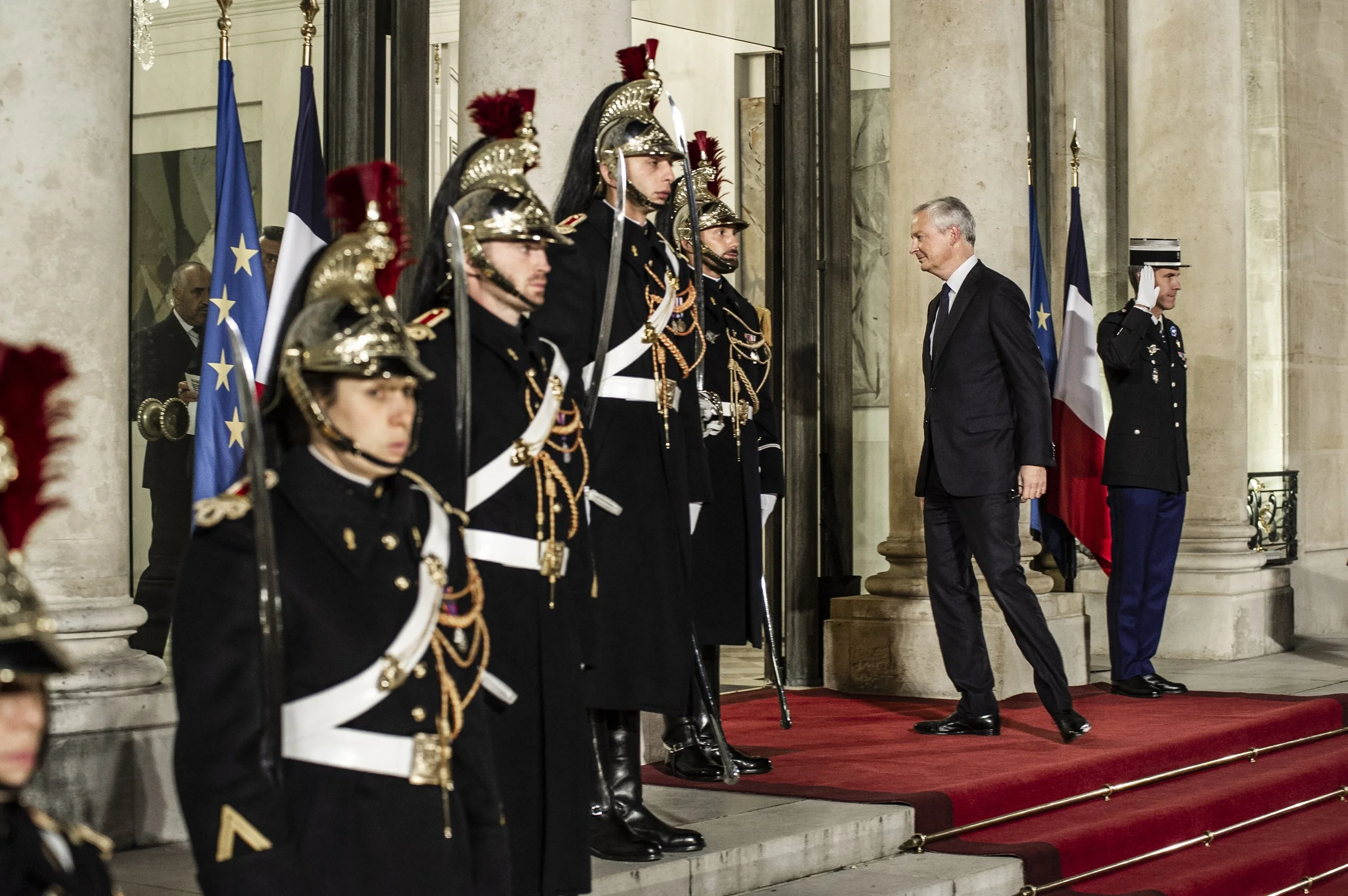 Un homme en costume sombre est en train de saluer une rangée de soldats en uniformes traditionnels avec chapeaux à plumes, lors d'une cérémonie officielle dans un bâtiment élégant avec des drapeaux français en arrière-plan.
