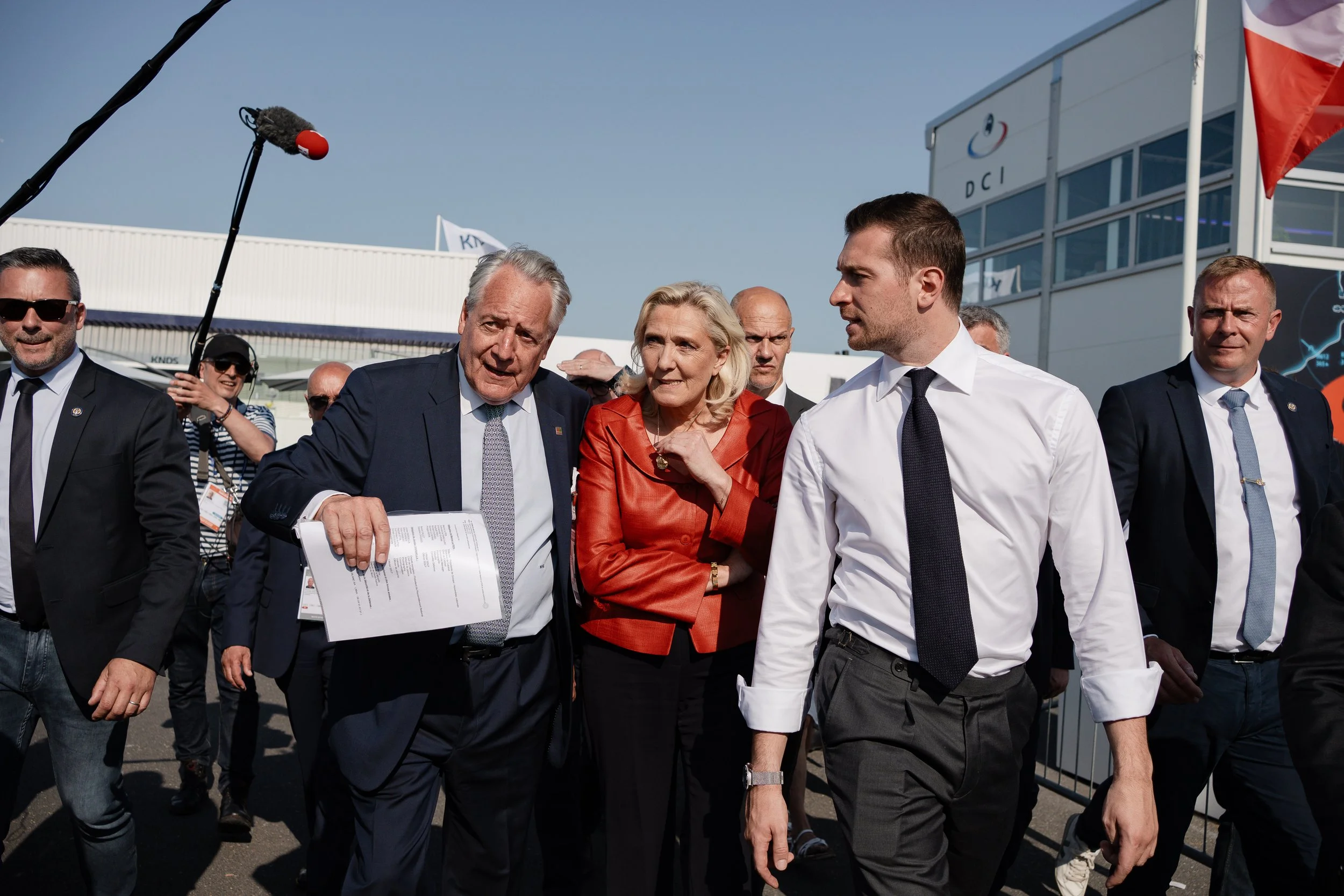 Groupe de personnes en costume discutant à l'extérieur lors d'un événement public, avec un bâtiment en arrière-plan.