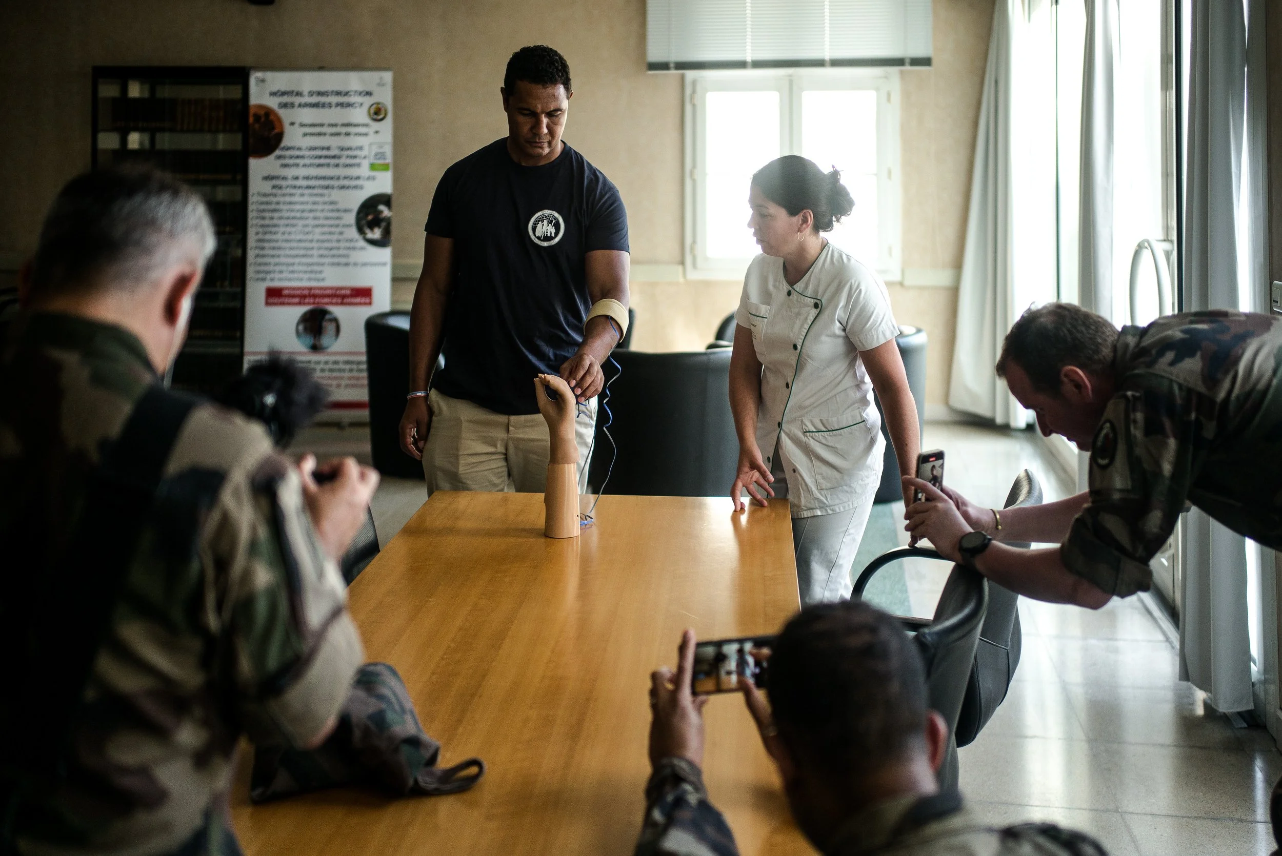 Un groupe de personnes, dont certains en uniformes militaires, observe un mannequin de bras humain avec des électrodes, sous la supervision d'une femme en uniforme médical, dans une salle bien éclairée.