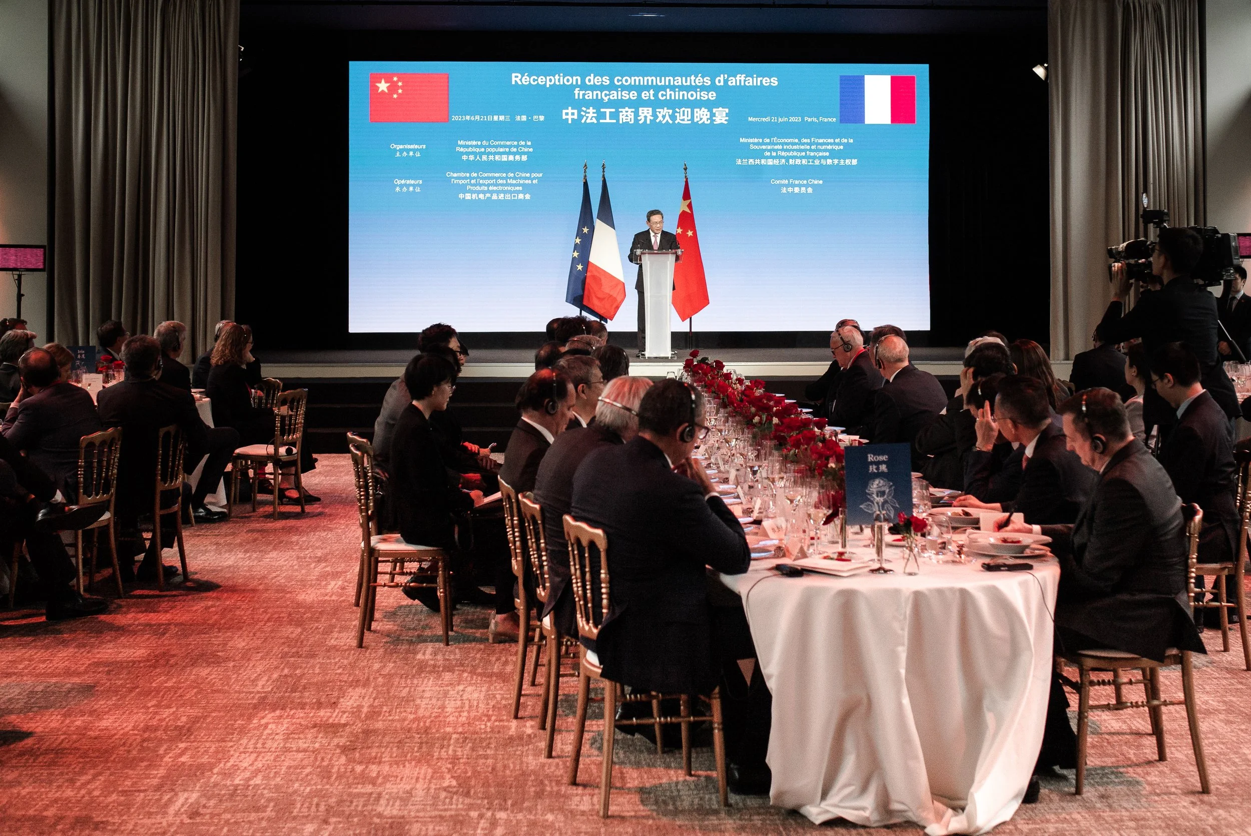 Une cérémonie avec des personnes assises autour d'une longue table, avec un écran géant derrière avec des drapeaux de la Chine et de la France, et un homme parlant à un podium.