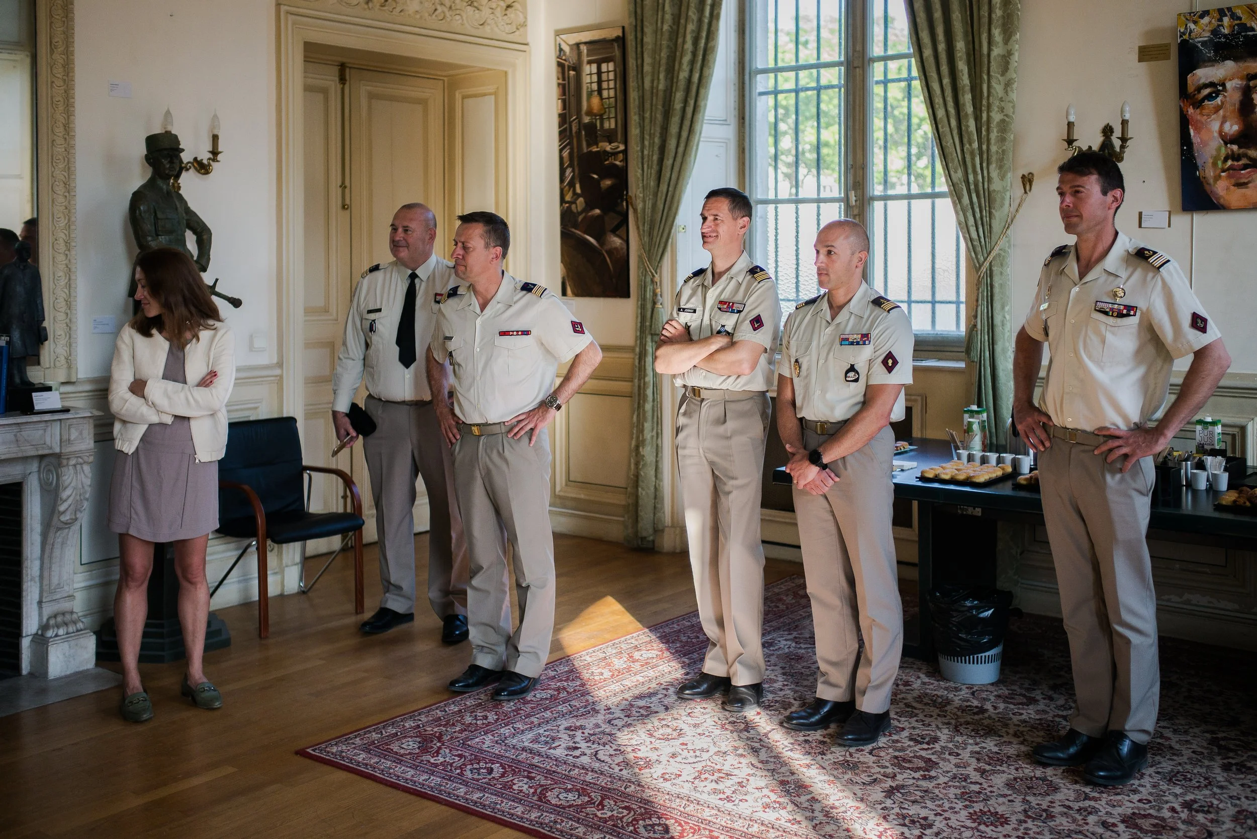 Une femme et cinq hommes en uniforme militaire se tiennent dans une pièce élégante avec chaises, tableaux et rideaux verts, dans une posture de prise de parole ou de présentation.