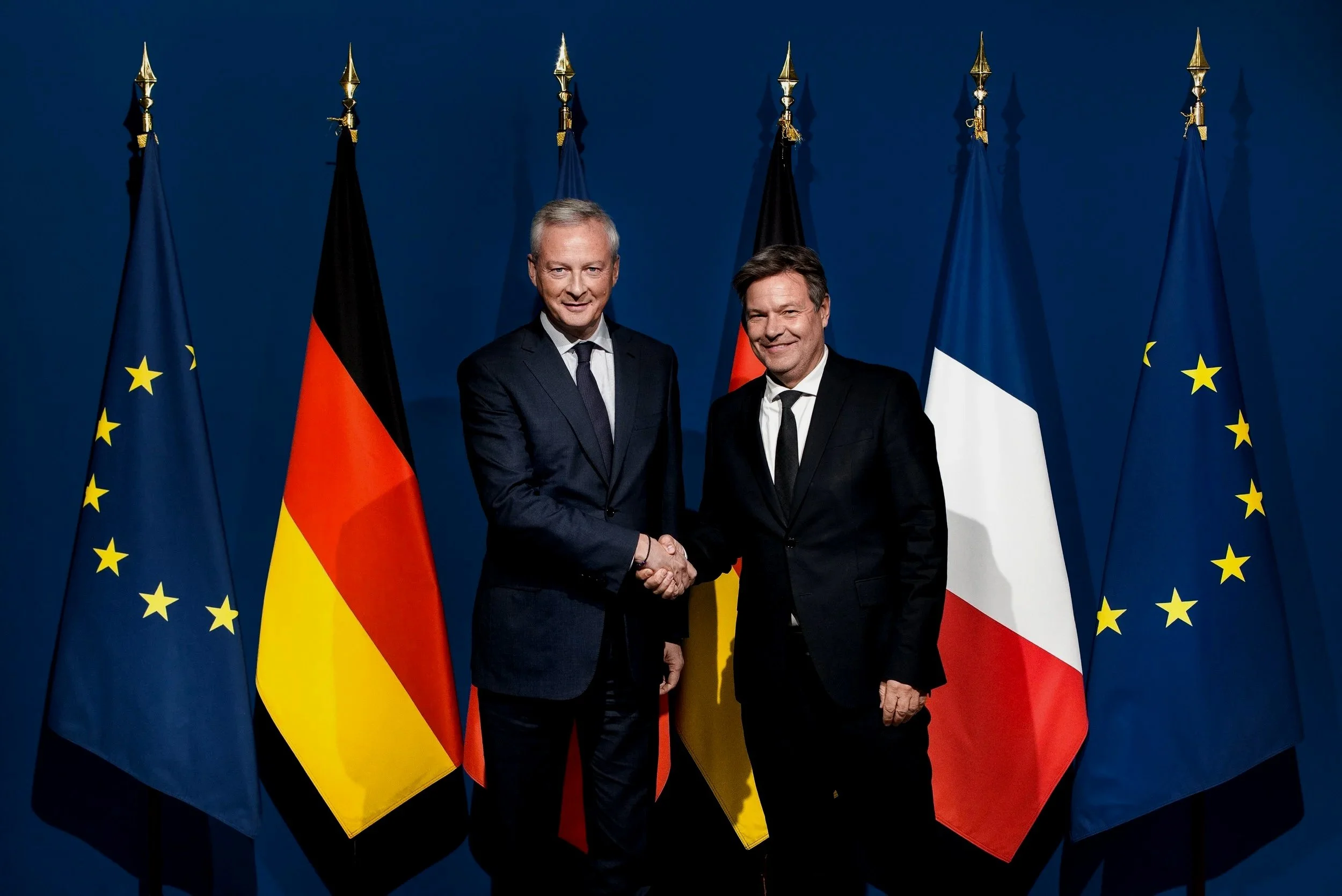 Deux hommes en costume se serrent la main devant un fond de drapeaux européens, français, allemands et italiens.
