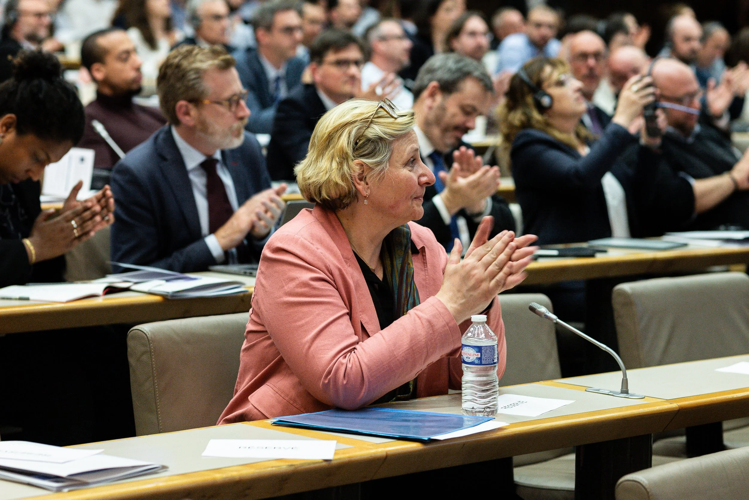 Une femme assise à une table lors d'une conférence, applaudissant, avec une bouteille d'eau devant elle et un microphone. Plusieurs personnes en arrière-plan aussi applaudissent, dans une salle de conférence formelle.