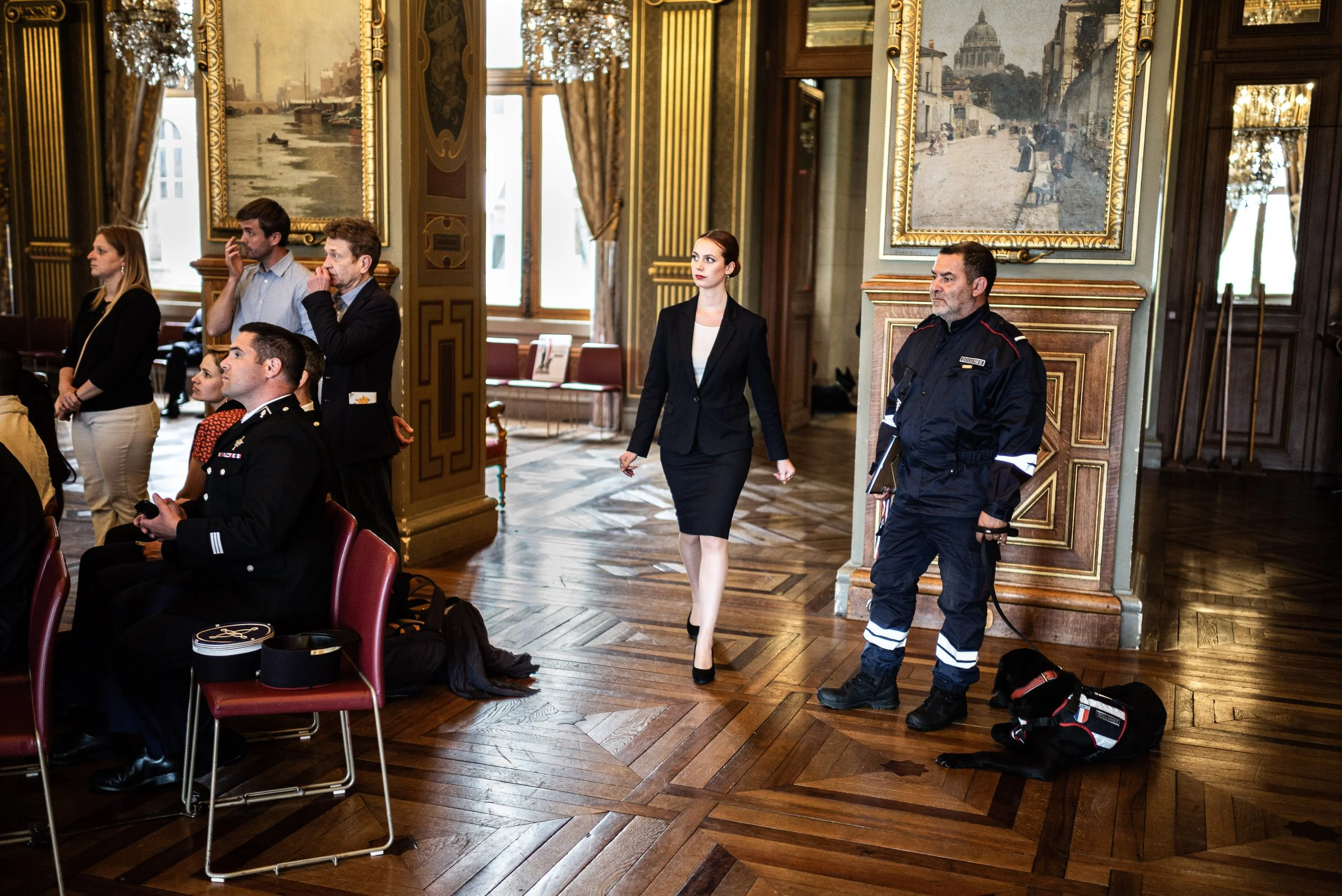 Une salle élégante avec plusieurs personnes, dont une femme en costume noir marchant, un officier de police, et d'autres personnes assises ou debout. La pièce est décorée avec des peintures, des chandeliers, et un parquet en bois.