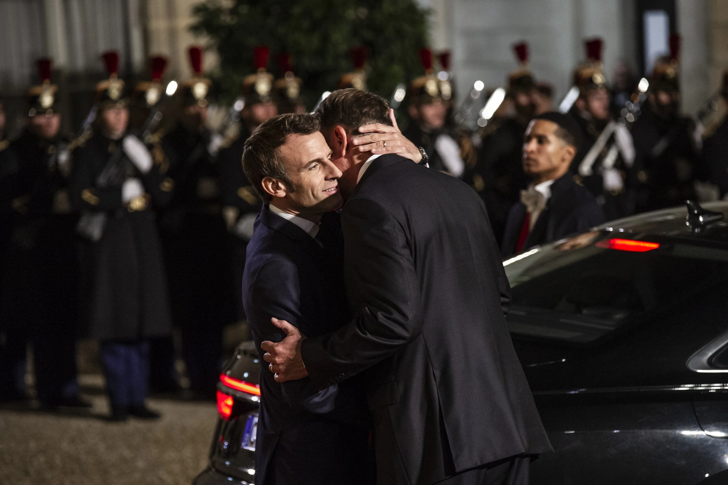 Deux hommes en costume qui s'embrassent lors d'une cérémonie, avec une rangée de militaires en uniforme en arrière-plan