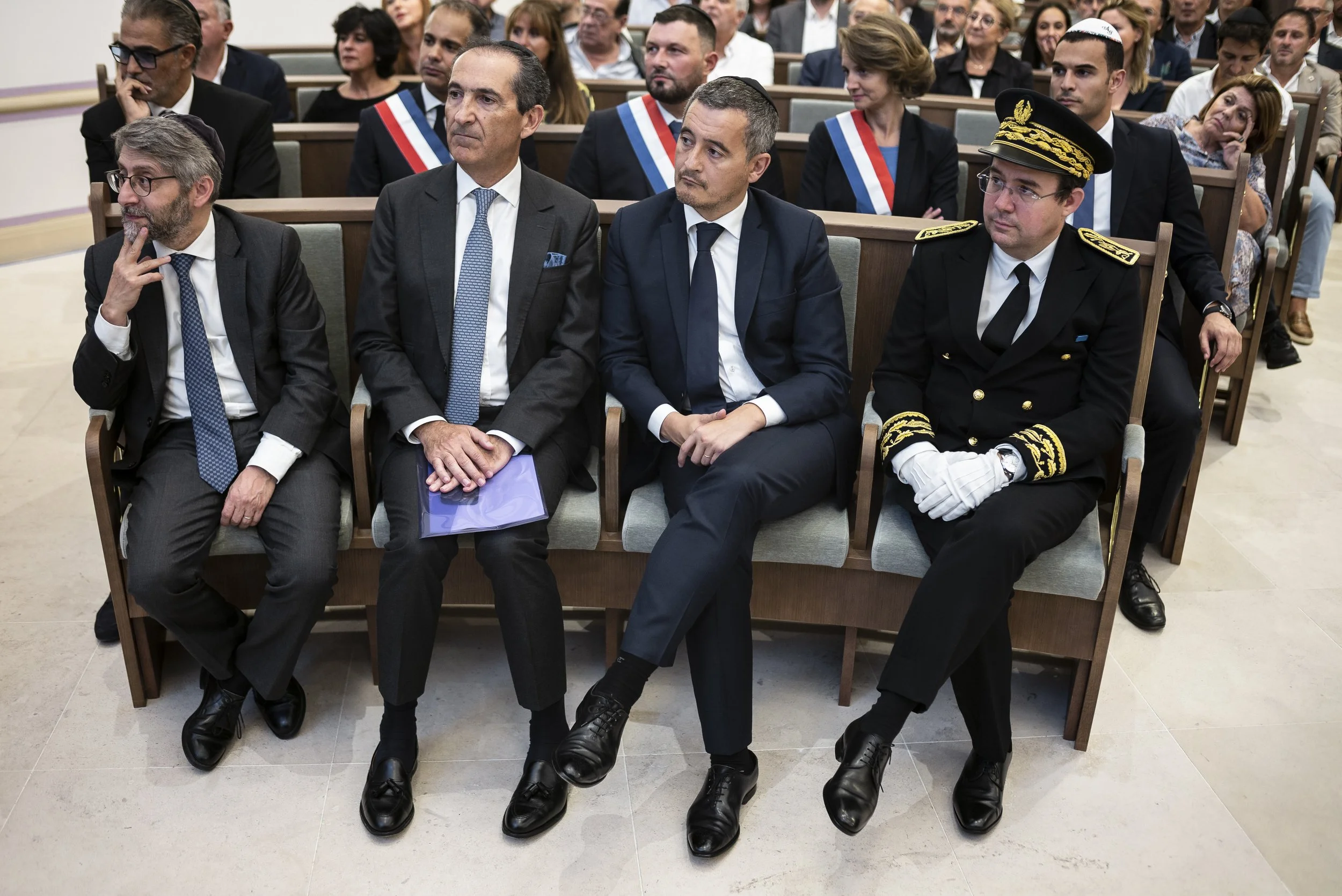 Des personnes assises en rangée lors d'une cérémonie officielle, parmi lesquelles un homme en uniforme militaire et d'autres en costumes, portant des écharpes tricolores.