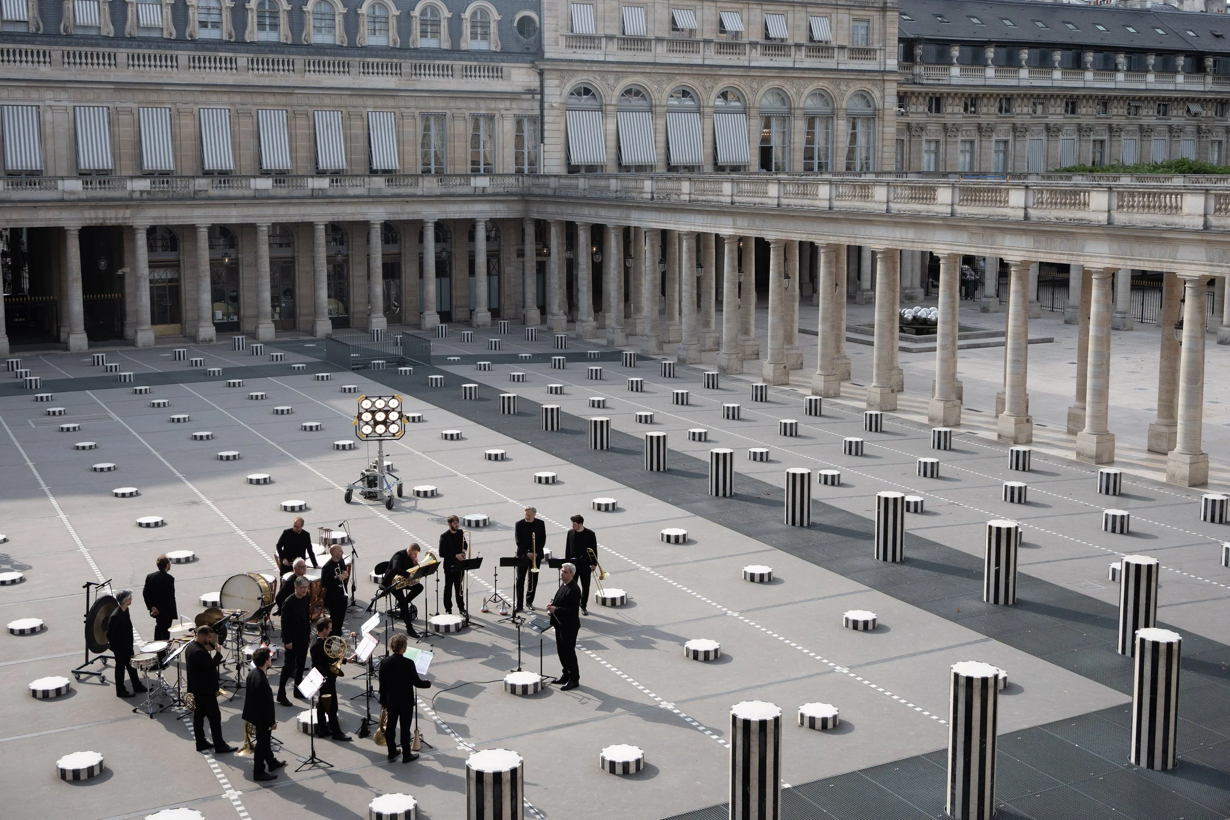 Un groupe de musiciens jouant dans une cour en plein air, entourée de colonnes et d'un bâtiment historique avec des fenêtres à volets, lors d'une répétition ou d'un concert dans un lieu architectural.