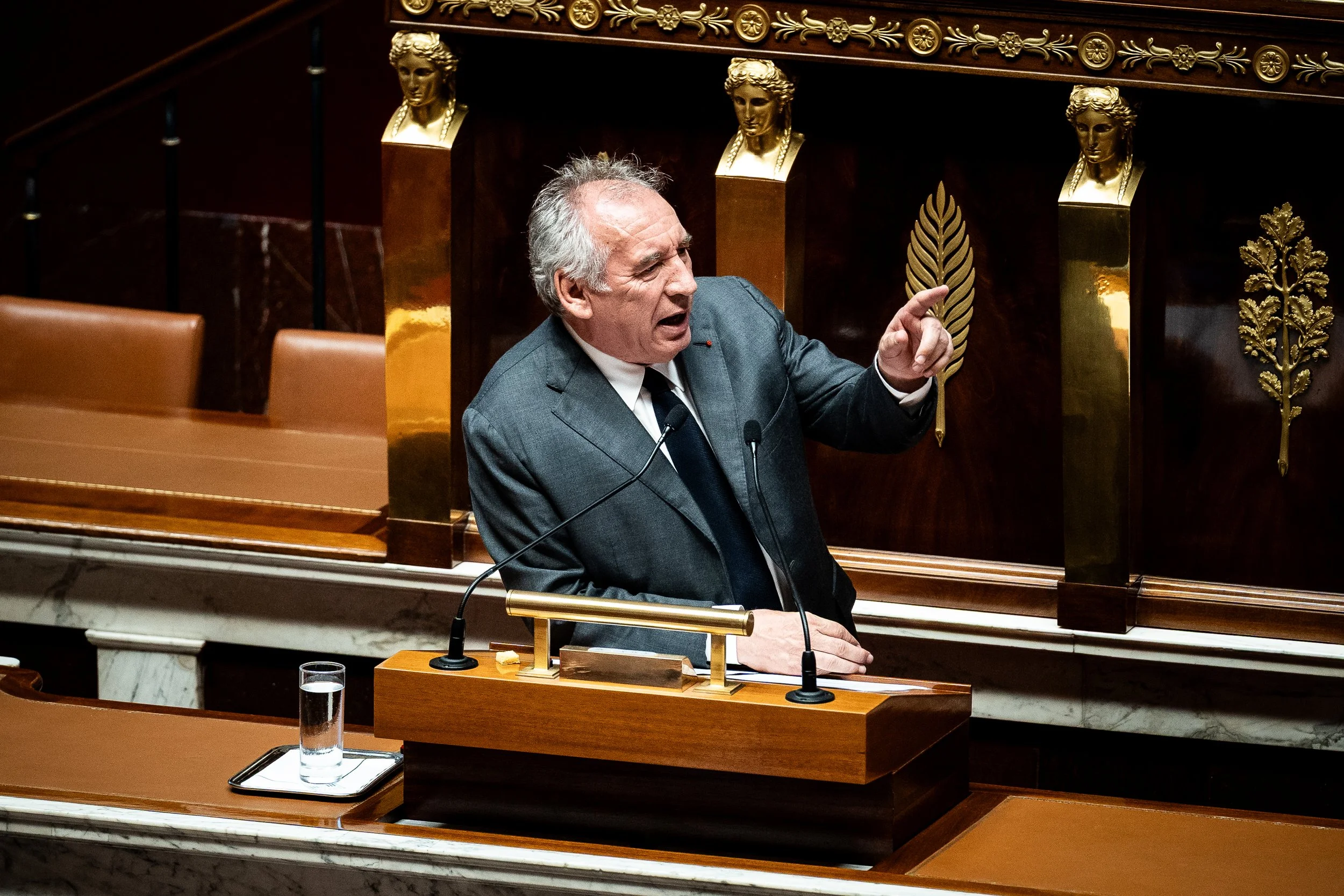 Un homme en costume parle à la tribune dans une salle ornée de décorations en or, avec des bustes en or et des motifs végétaux en arrière-plan.
