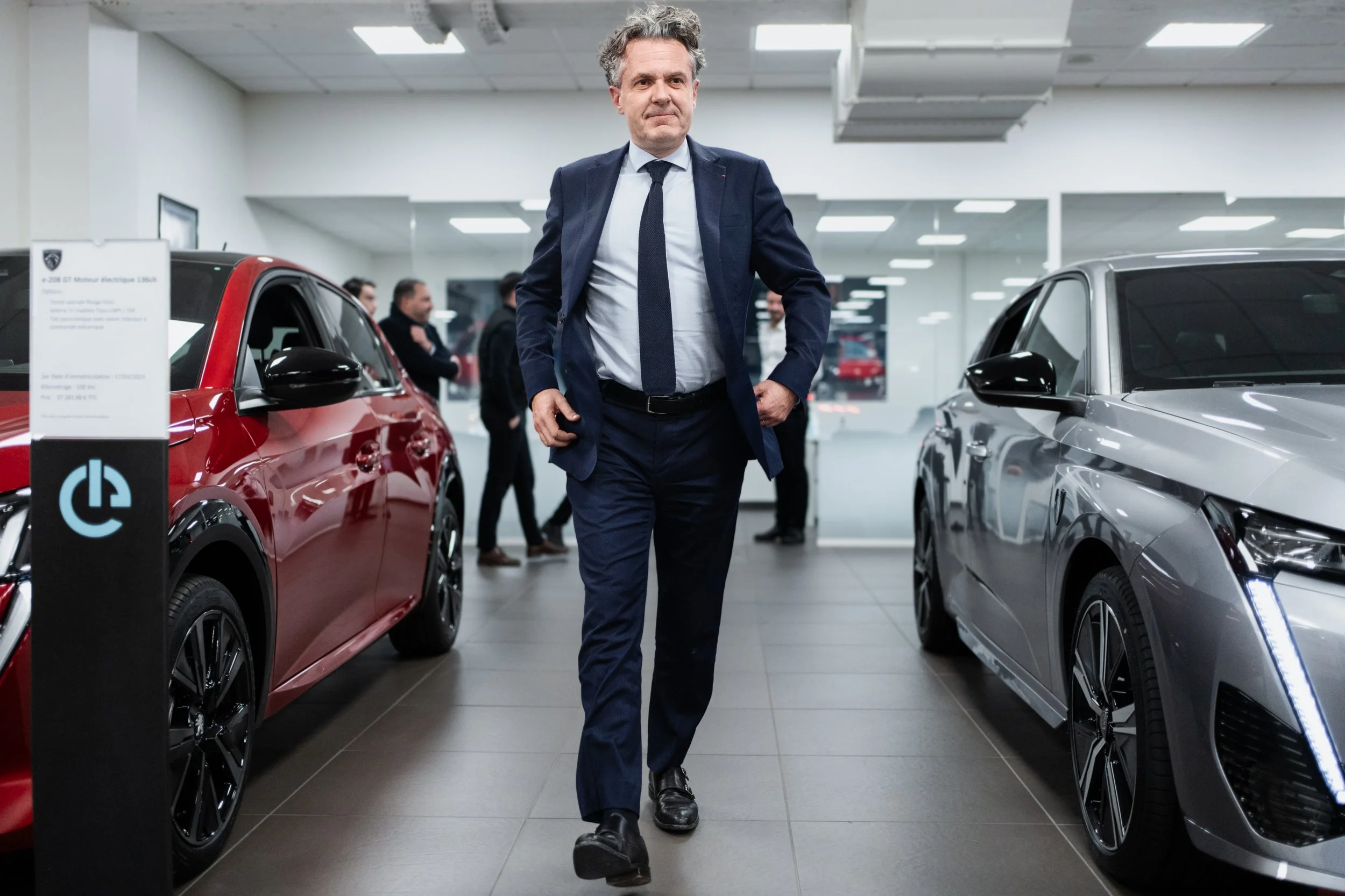 Un homme en costume se promène entre deux voitures dans un salon automobile, avec plusieurs autres personnes en arrière-plan.