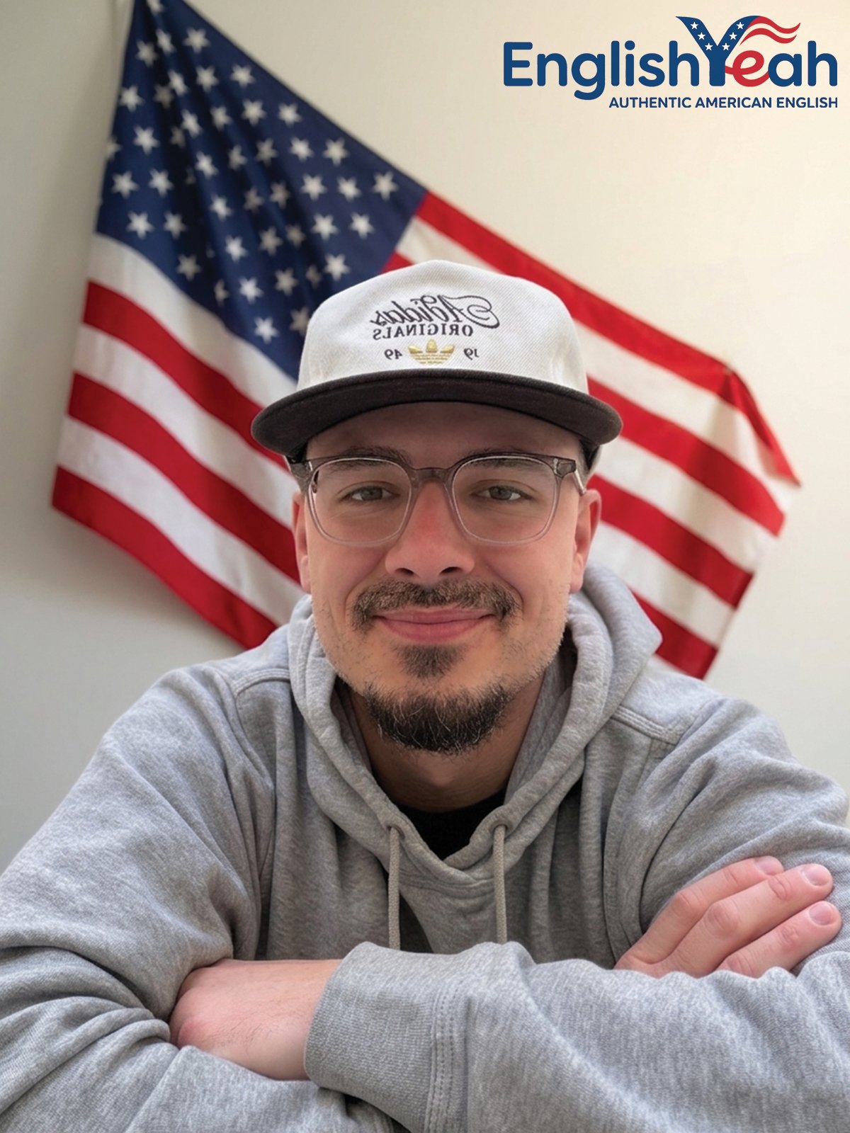 A smiling man wearing glasses and a gray hoodie, sitting with arms crossed, in front of an American flag hanging on a wall. The top right corner displays the logo 'English Yeah' with the slogan 'Authentic American English'.