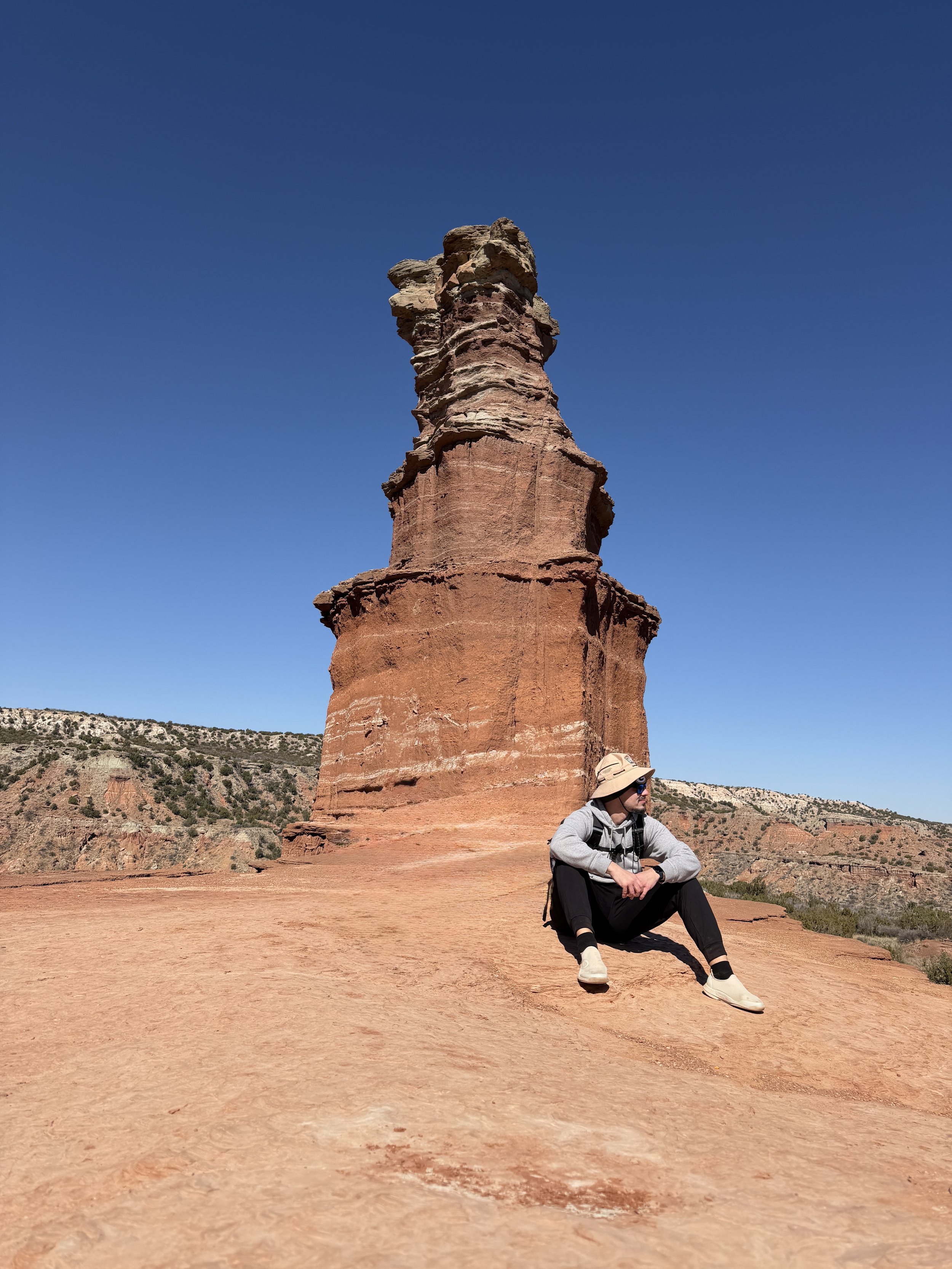 A person wearing a hat, sunglasses, and casual clothes sitting on red rock in a desert landscape with a large rock formation behind them and a clear blue sky above.