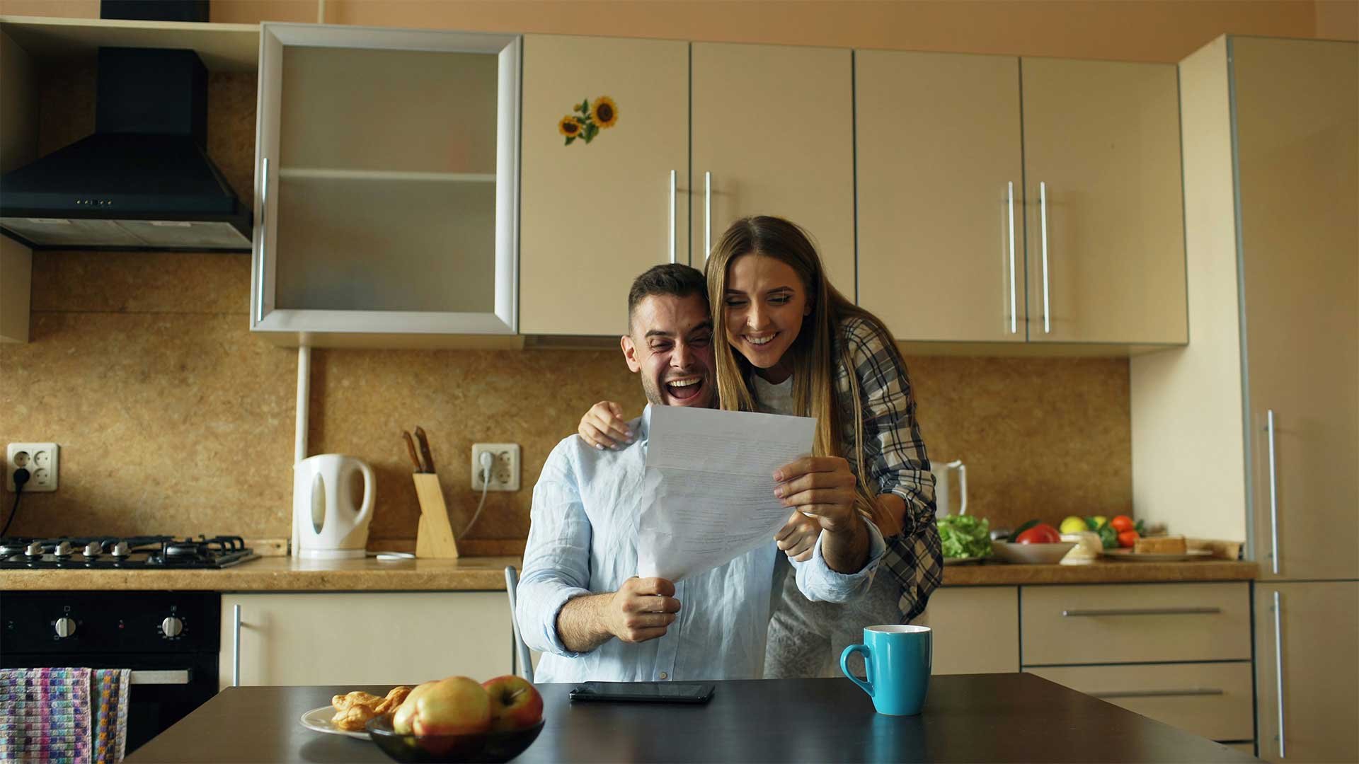 Smiling couple reviewing a document together in a kitchen, reacting with excitement as they read the paper