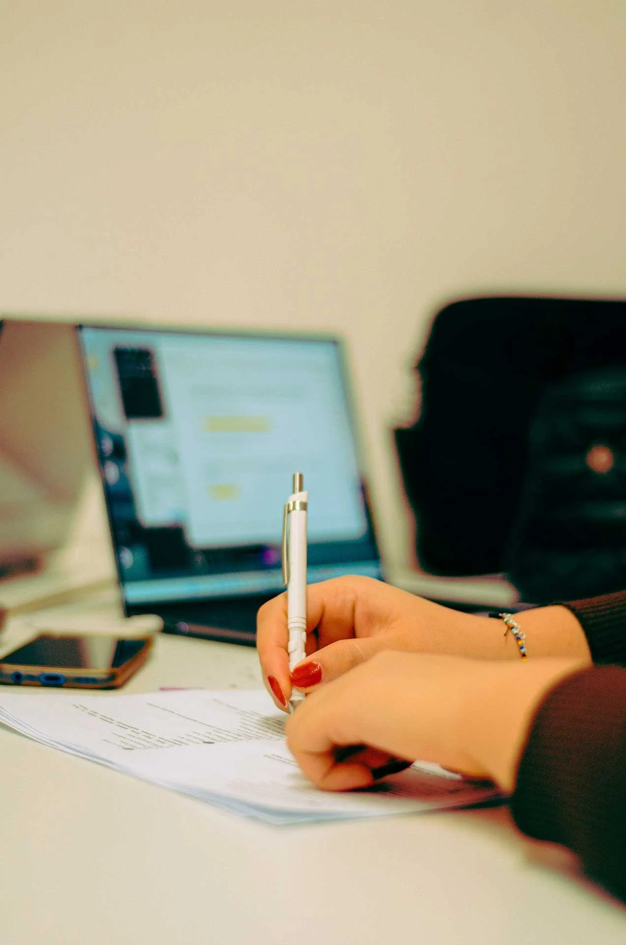 Close-up of a person's hand as they fill out paperwork by hand, with a laptop open in the background