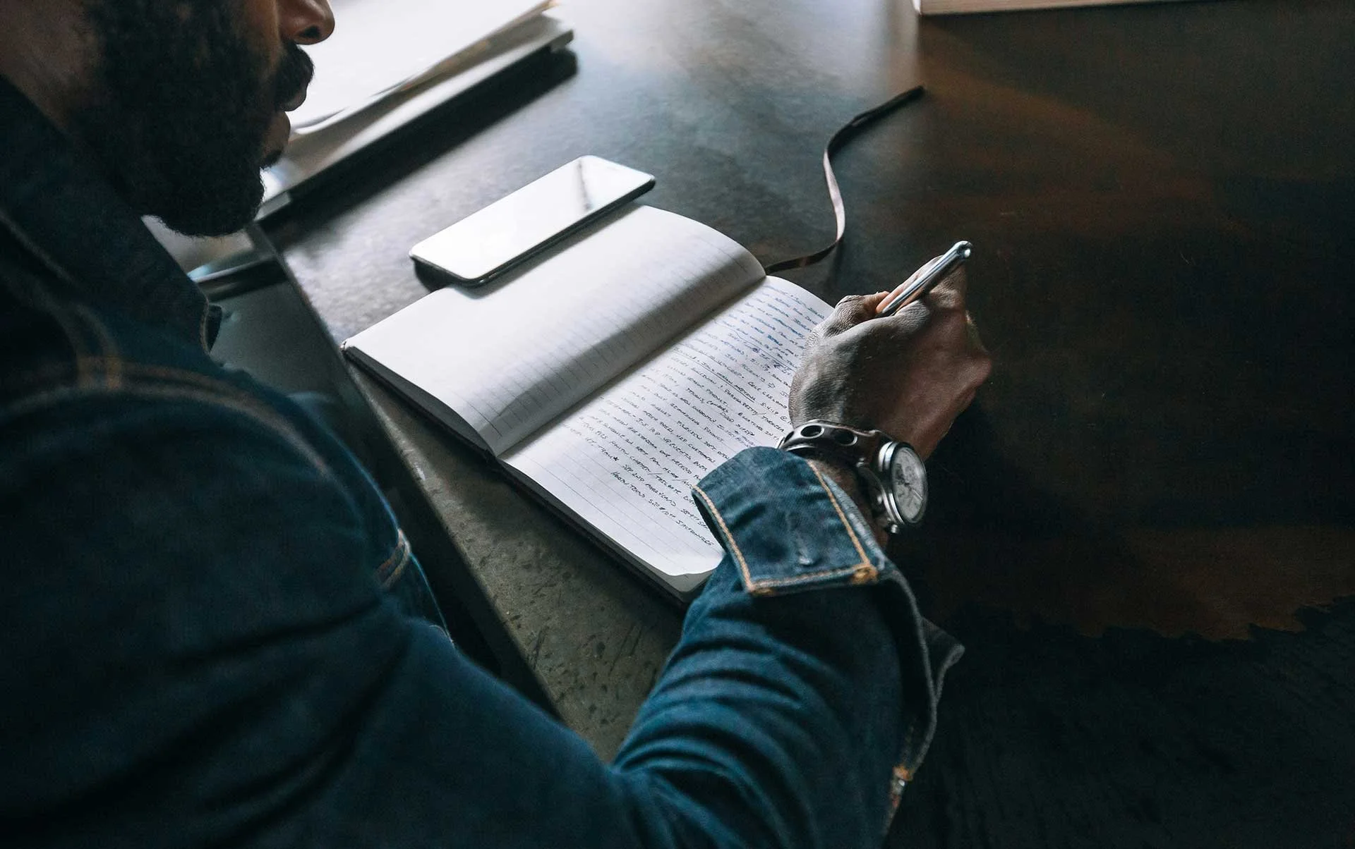 Man writing in a notebook at a desk with a smartphone nearby, focused on planning and note-taking