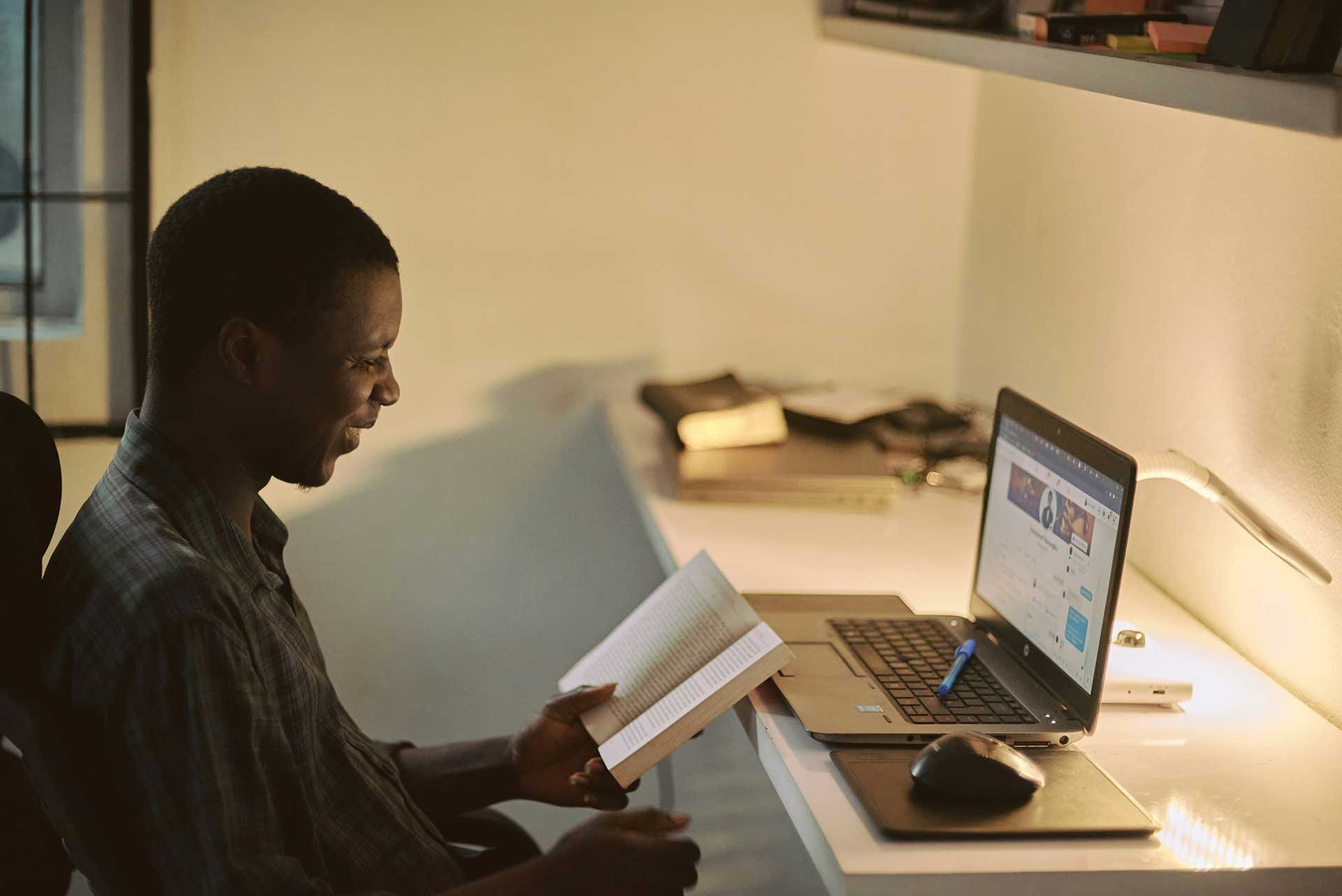 Man reading a book at a desk with a warm light, smiling as he reviews the page