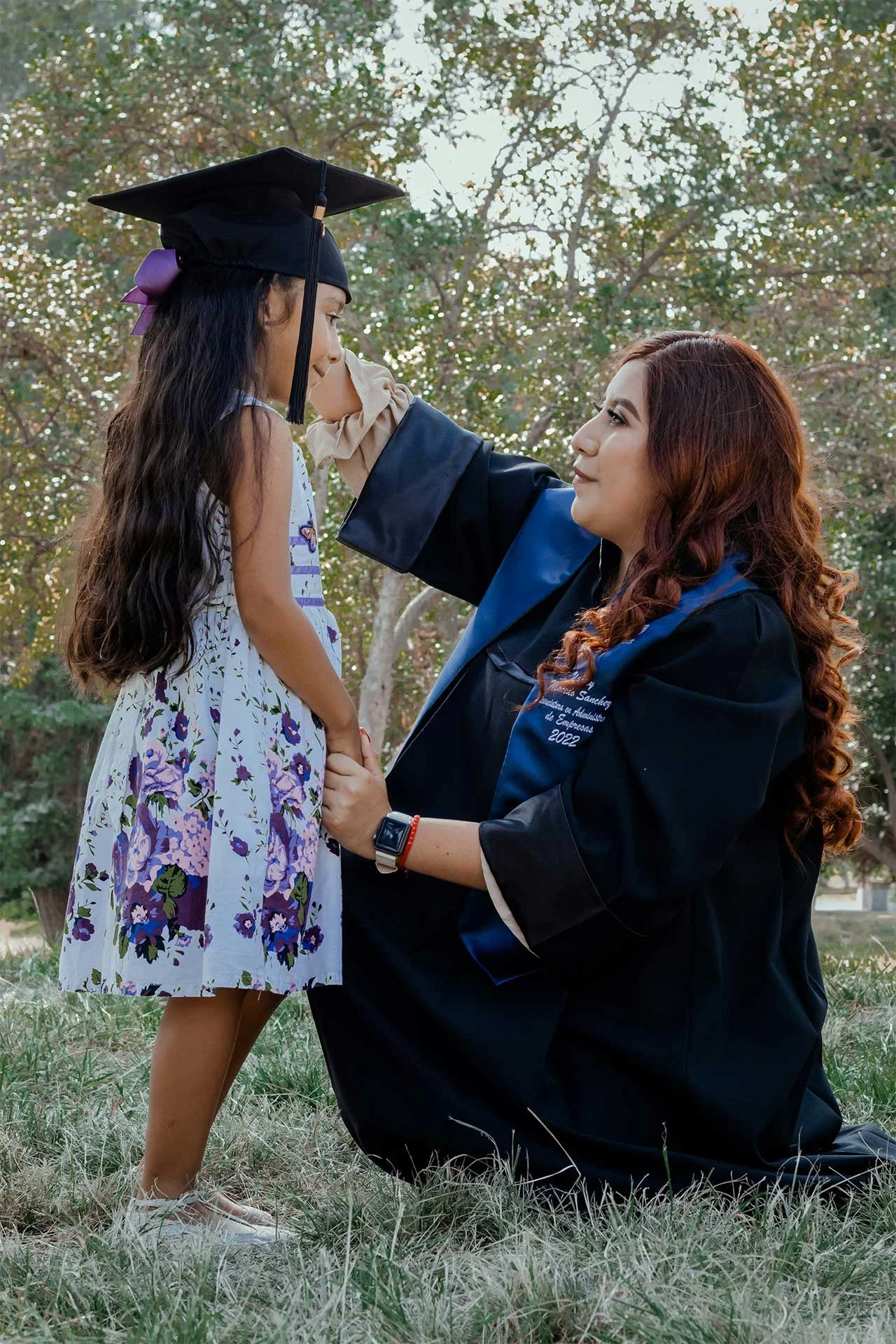 Adult college graduate kneeling down to adjust her graduation cap on a child's head. The two are softly smiling at each other in a proud, supportive moment.