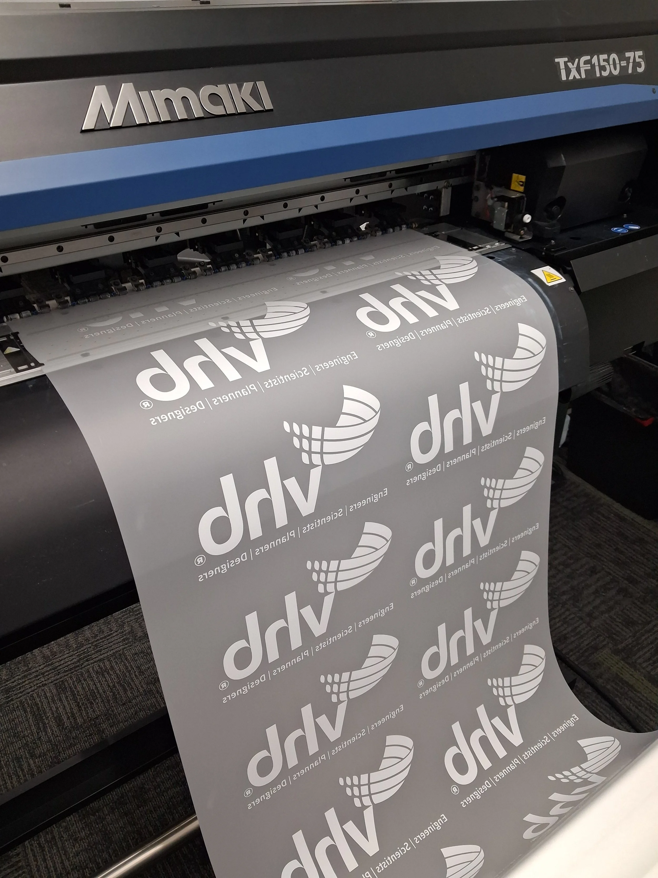 A large dtf printer printing a sheet with the logo 'VHB' and the words 'Engineers | Scientist | Planners | Designers' repeated on it.