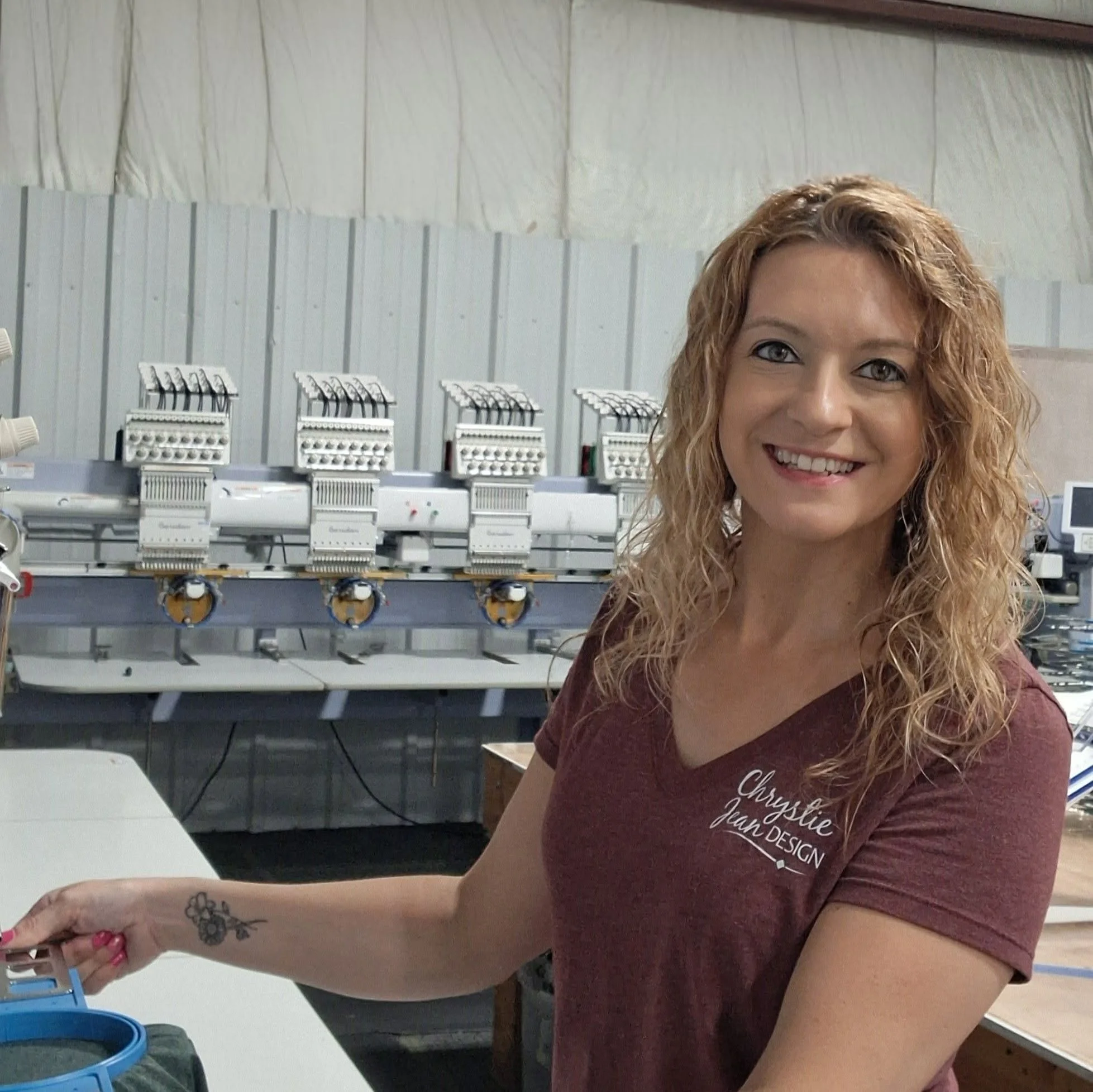 A woman with curly hair smiling in a room with laundry machines or embroidery machines in the background.
