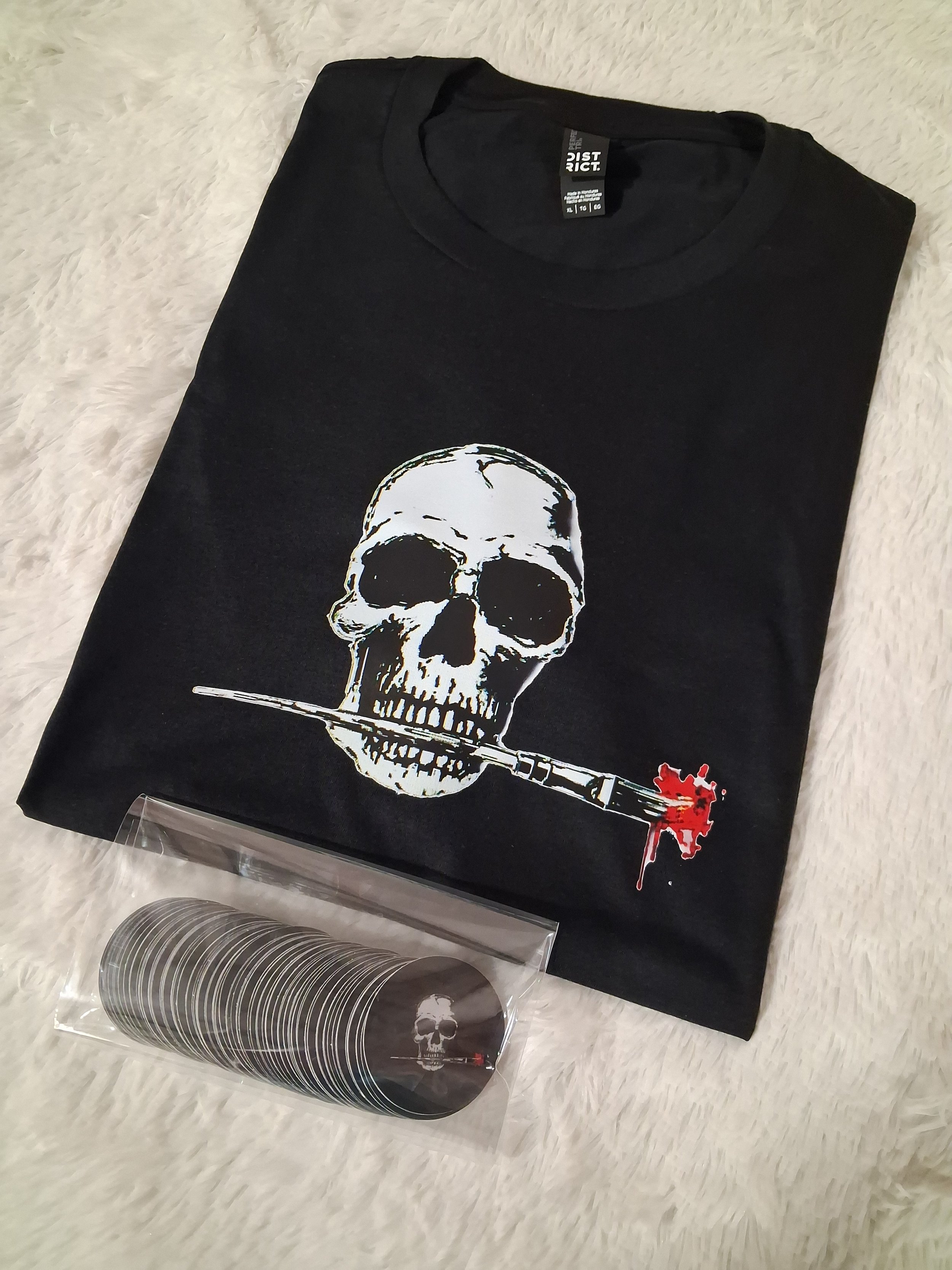 Black T-shirt with a white skull and a bleeding knife graphic, placed on a white furry surface. next to a vinyl record with a skull design in a plastic sleeve.