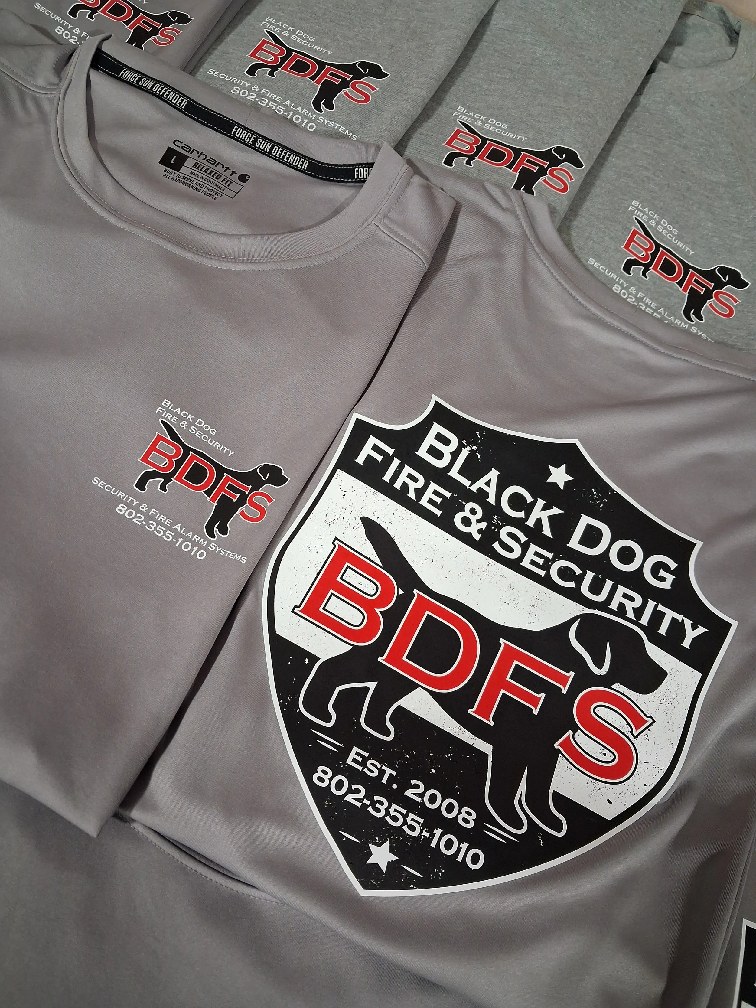 Gray t-shirt with a Black Dog Fire & Security logo and a shield emblem displaying the company name and contact information, alongside other gray t-shirts with the same logo in the background.